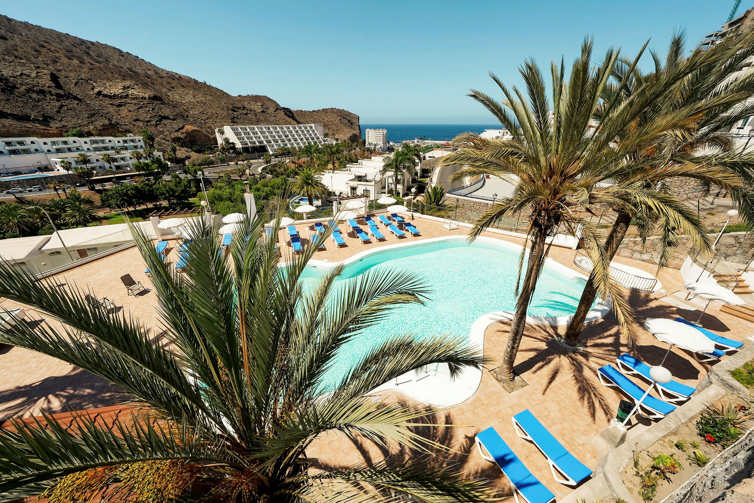 Udsigt over en hotelpool omgivet af liggestole og palmer med bjerge og hav i baggrunden. (AI-genereret tekst)