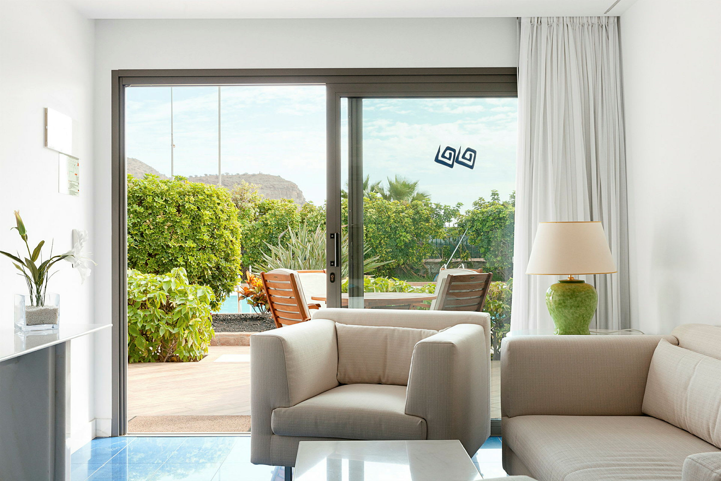 Billede af et rum med lys indretning, en lænestol og sofa, med en glasdør der fører ud til en terrasse med grønt. (AI-genereret tekst)