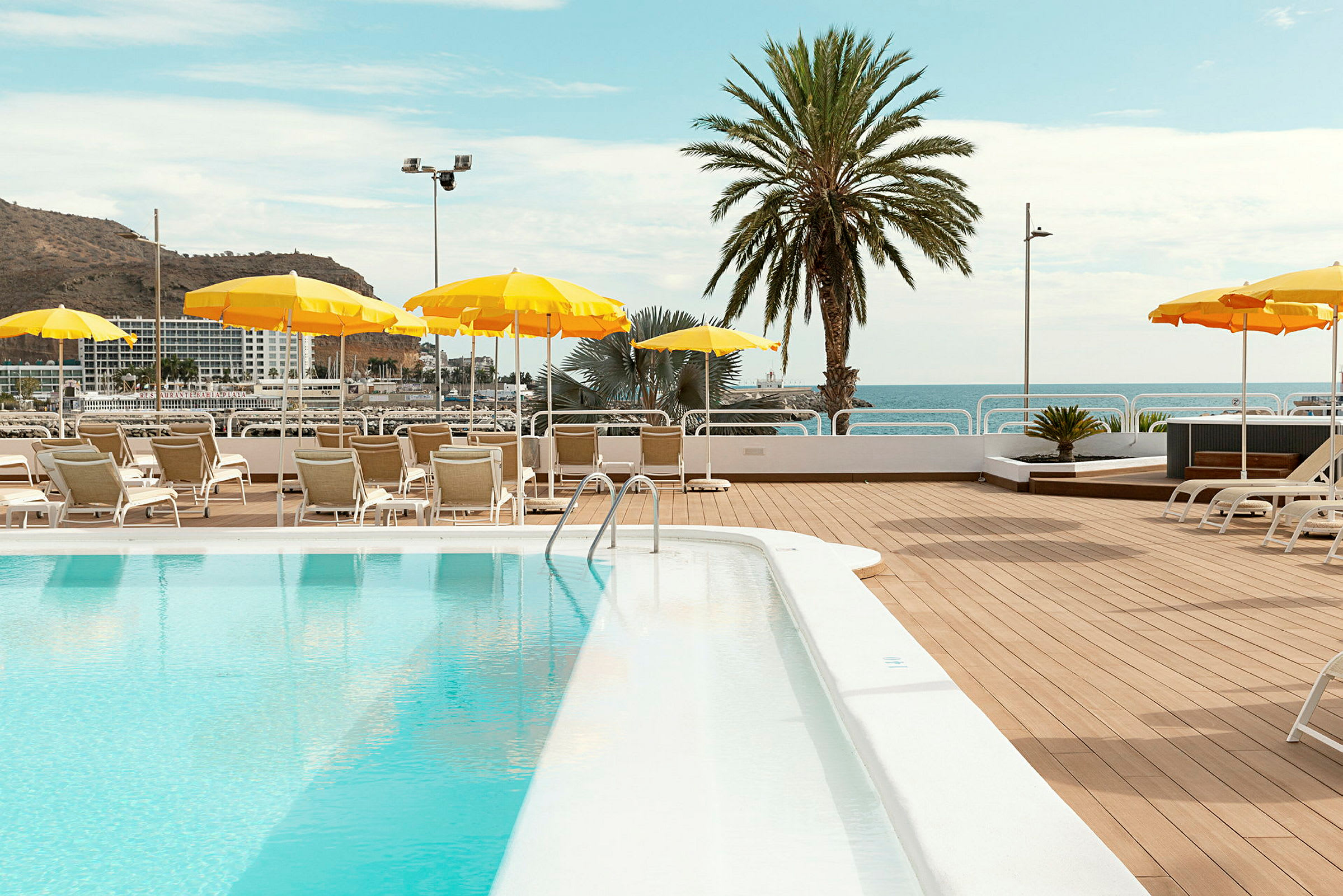 En solterrasse ved en pool med liggestole og gule parasoller placeret omkring. En palme og havet ses i baggrunden. (AI-genereret tekst)