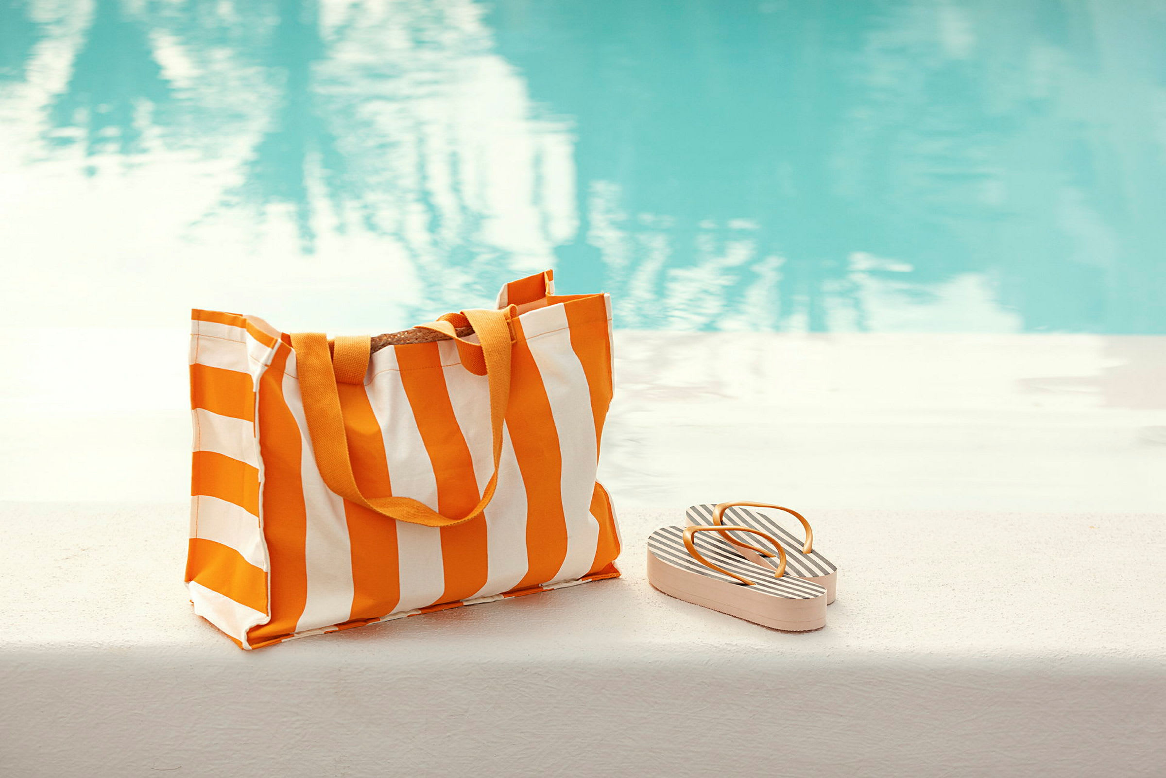 En stribet strandtaske og sandaler ligger ved siden af en pool. (AI-genereret tekst)