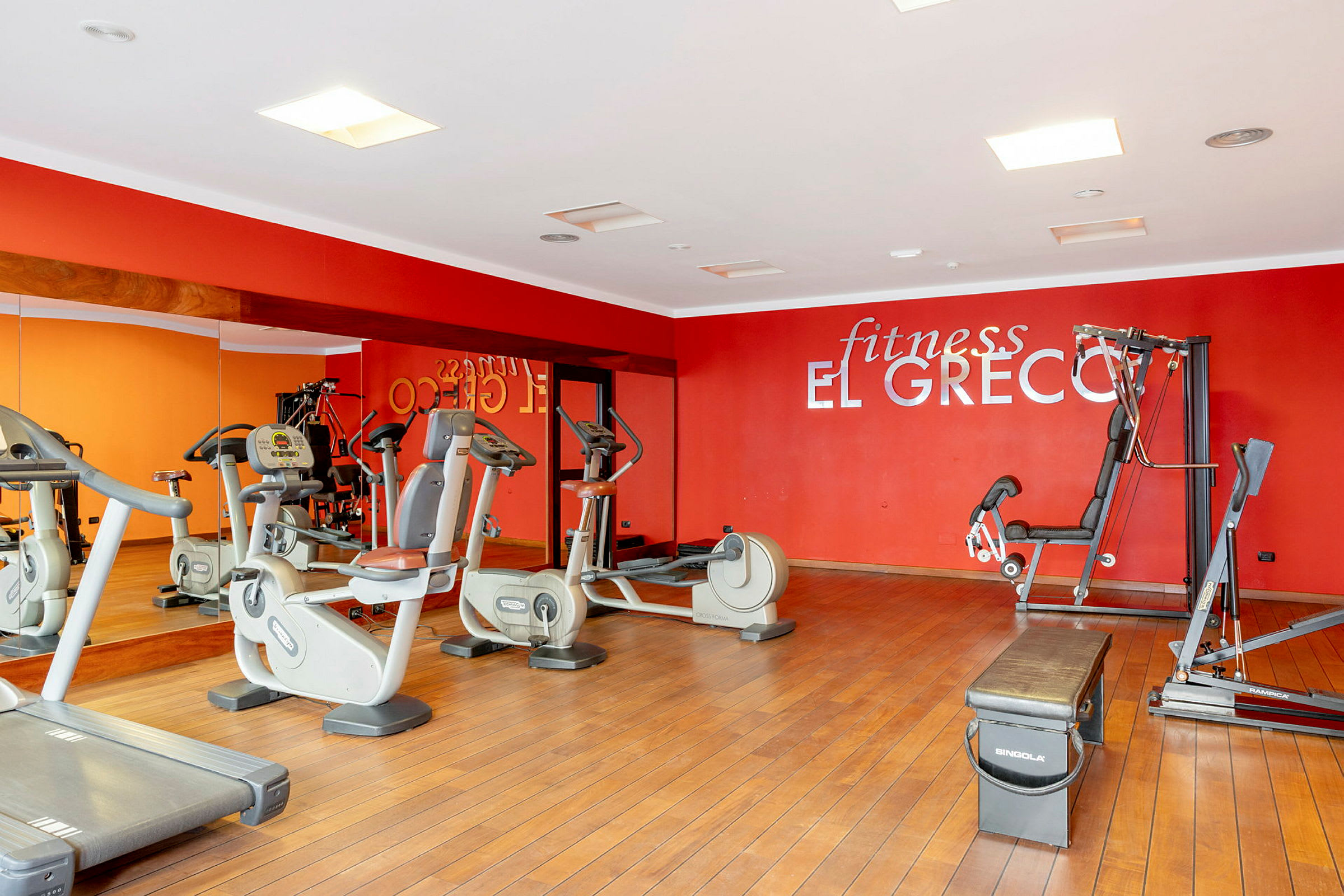 Et fitnesscenter med træningsmaskiner, spejle og en rød væg med teksten 'fitness El Greco'. (AI-genereret tekst)