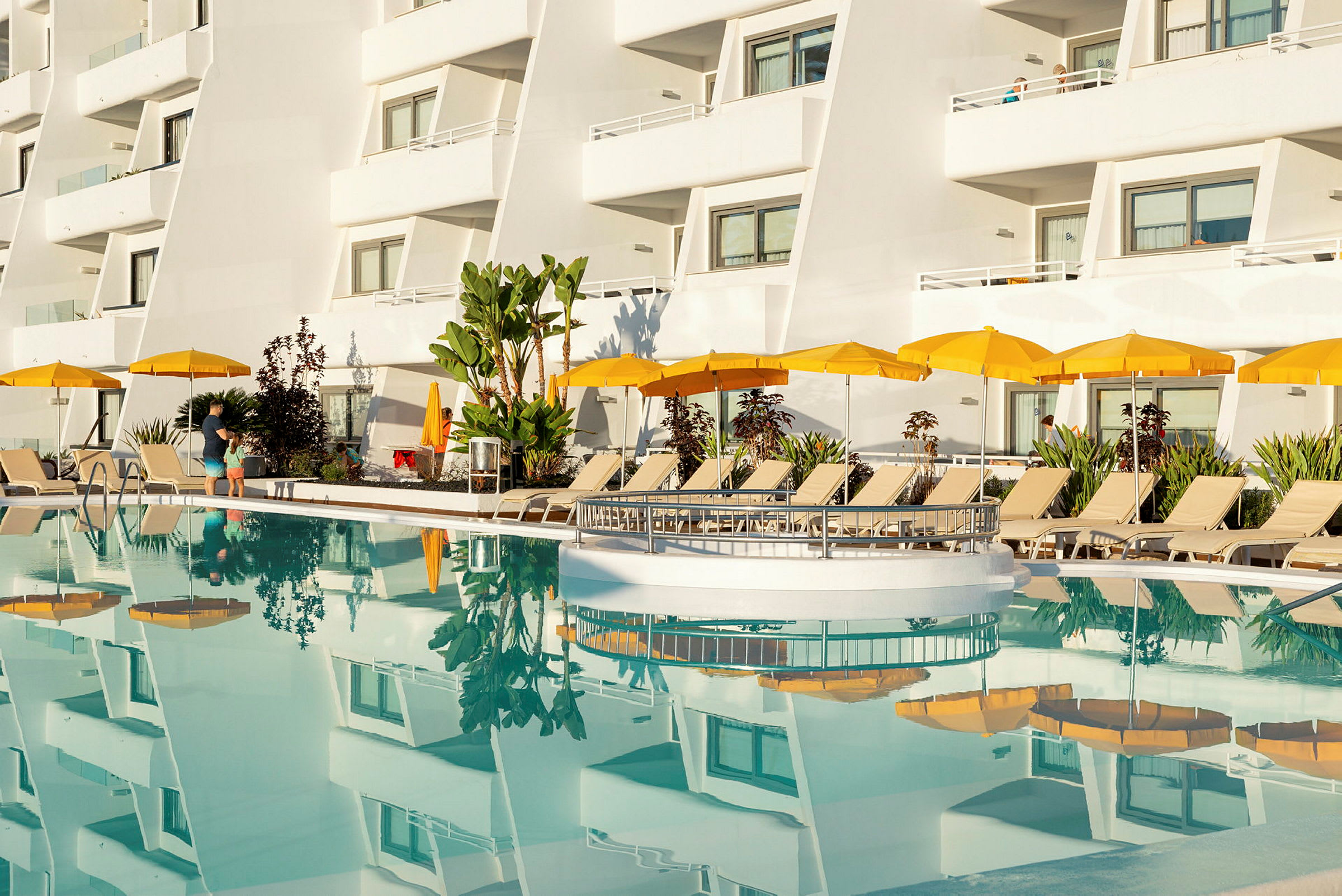 Hotellets facade med altaner og gule parasoller ved en pool. (AI-genereret tekst)
