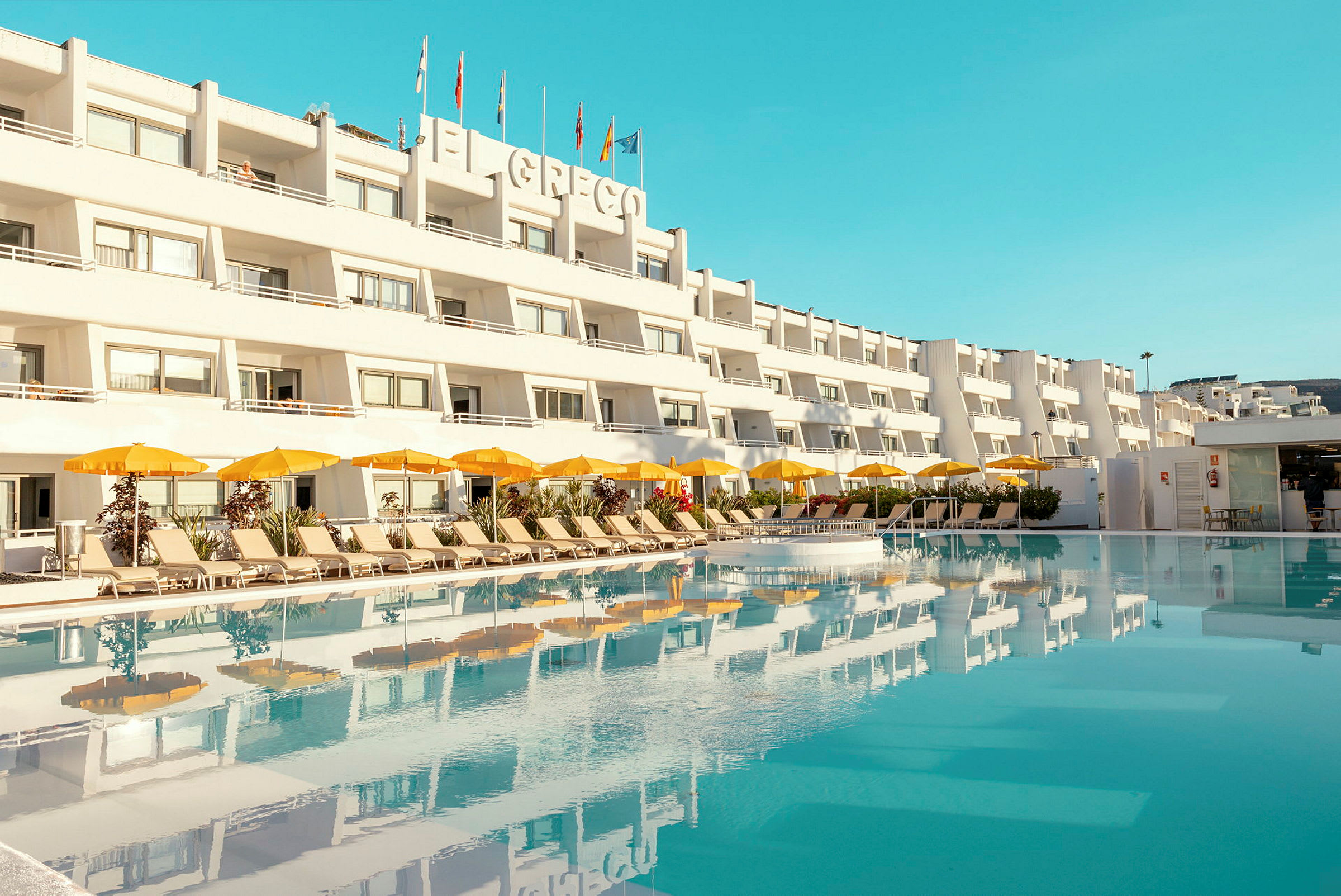 Hotelbygning med flere etager med en pool i forgrunden og solsenge samt gule parasoller. (AI-genereret tekst)