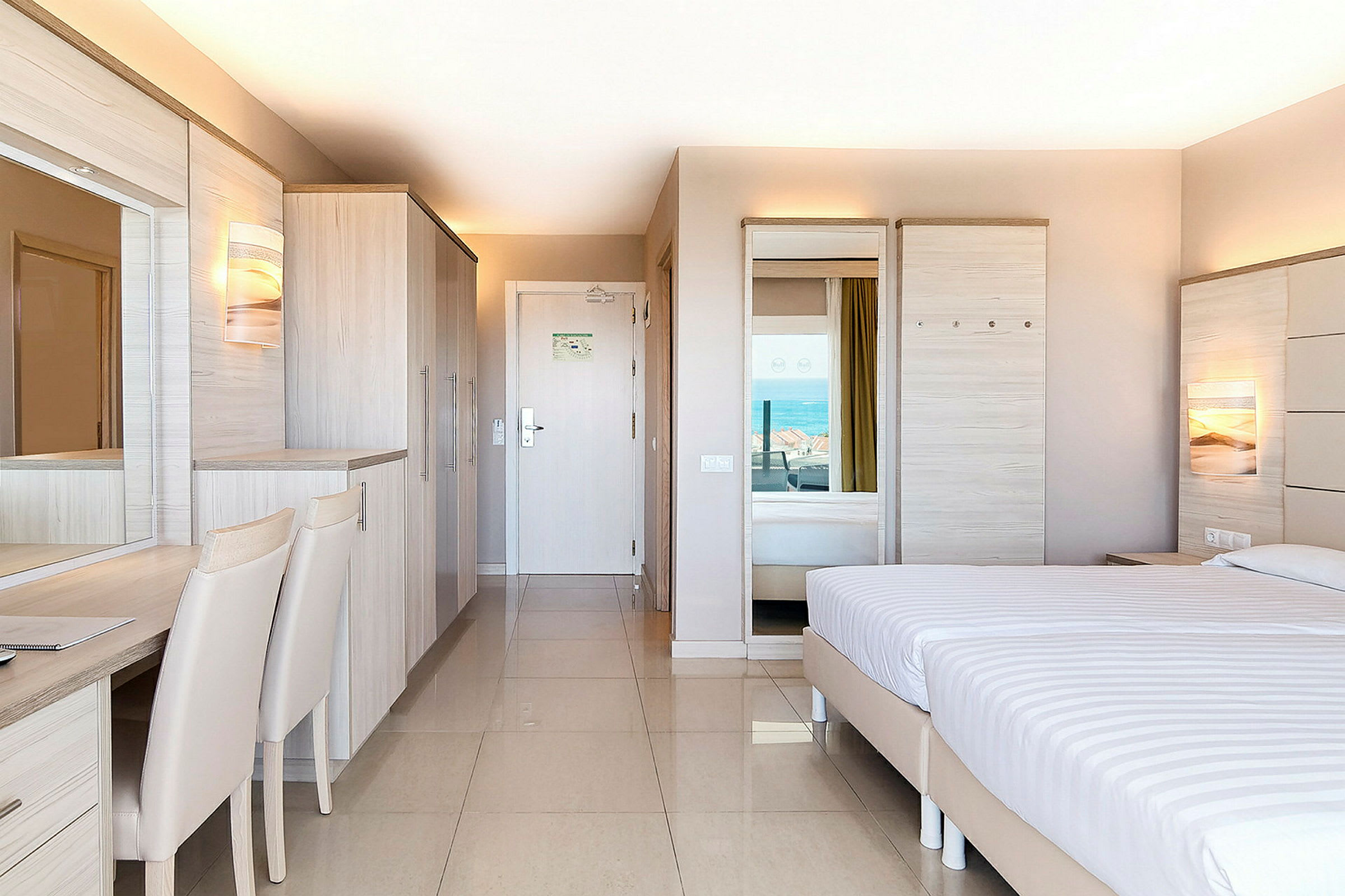 Et hotelværelse med moderne indretning, en dobbeltseng, garderobe og skrivebord, med udsigt til havet. (AI-genereret tekst)
