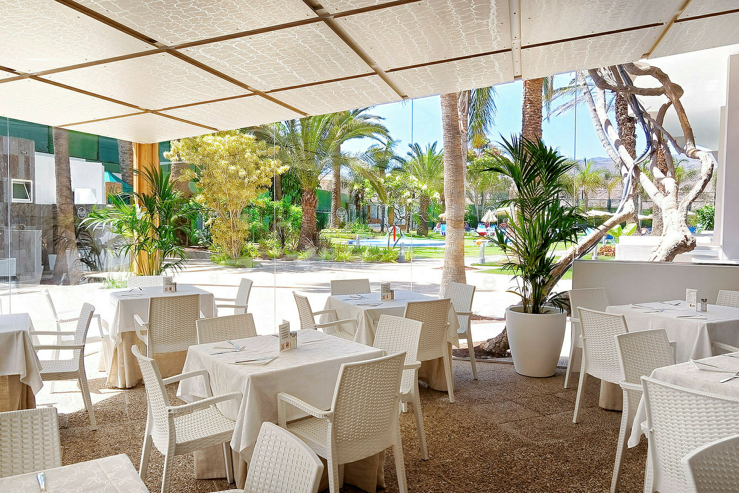 Udendørs restaurant med hvide borde og stole samt udsigt til en frodig have. (AI-genereret tekst)