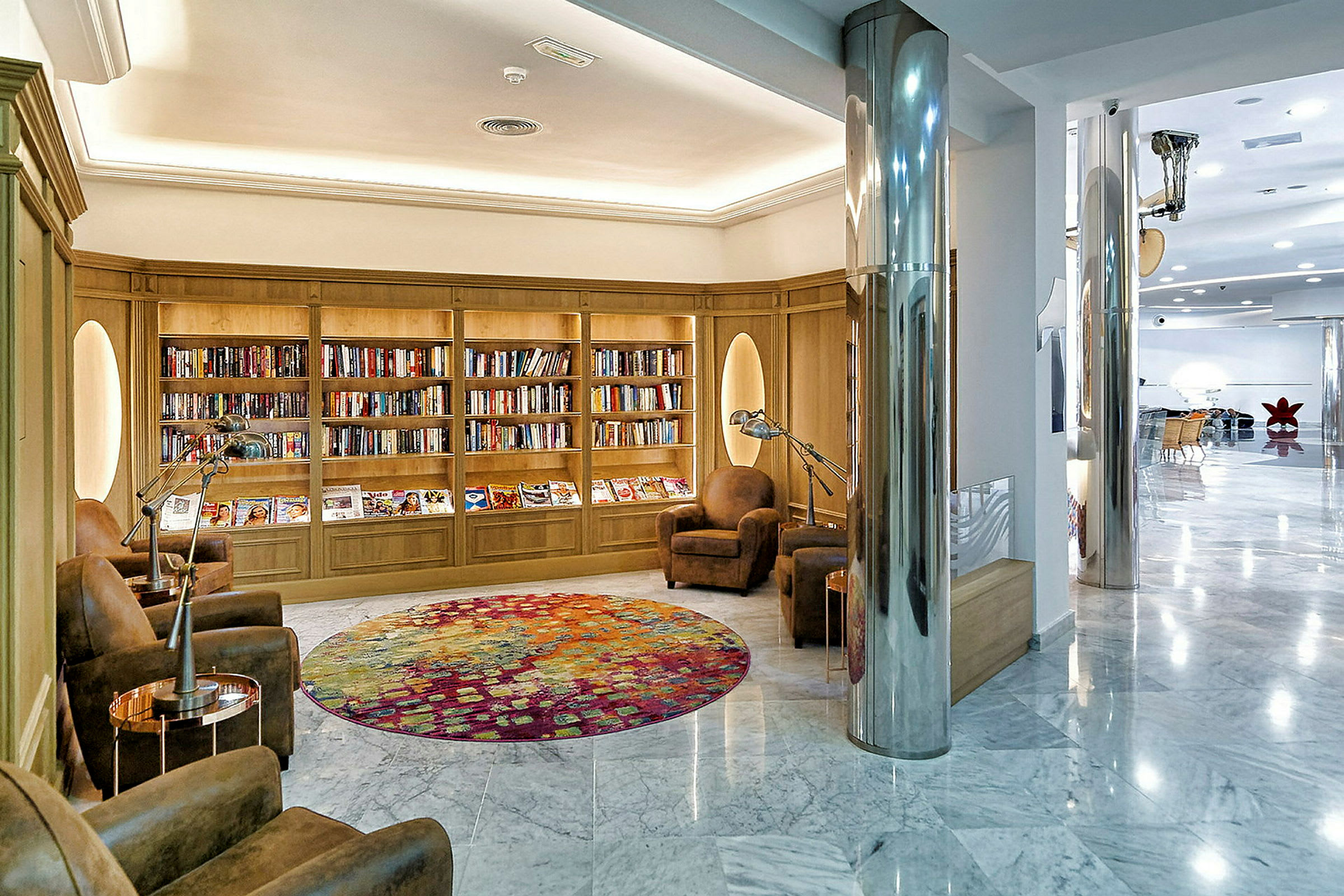 Et elegant hotelbibliotek med bogreoler, komfortable lænestole og et farverigt rundt gulvtæppe. (AI-genereret tekst)