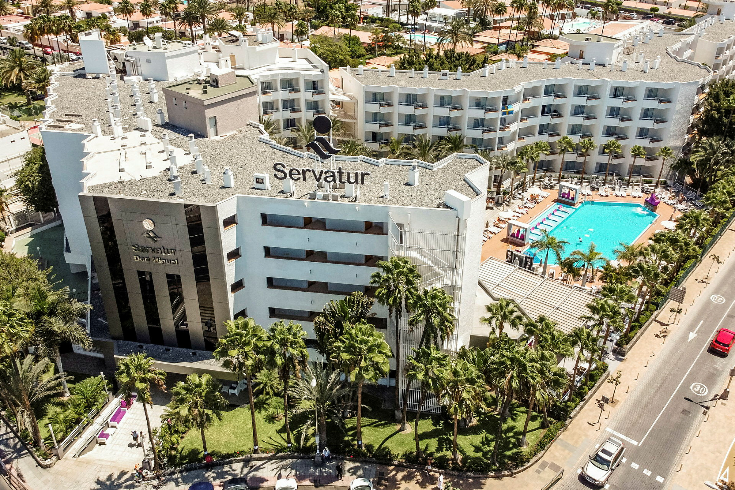 Luftfoto af hotelbygning med poolområde og omkringliggende grønne områder. (AI-genereret tekst)