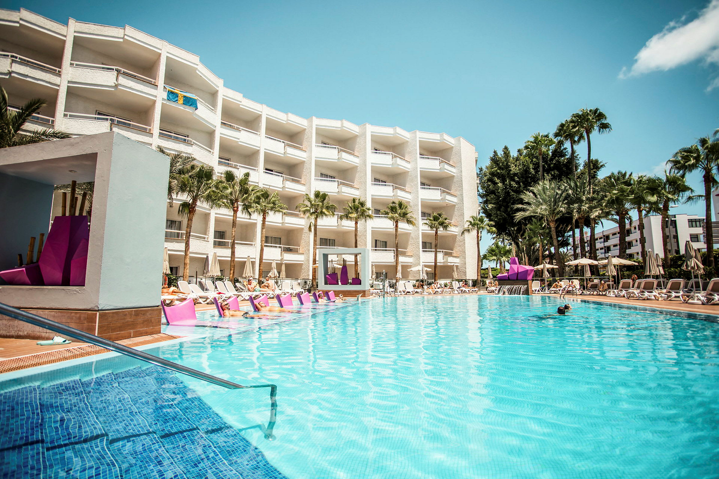 Hotelpool med liggestole og bygning i baggrunden. (AI-genereret tekst)