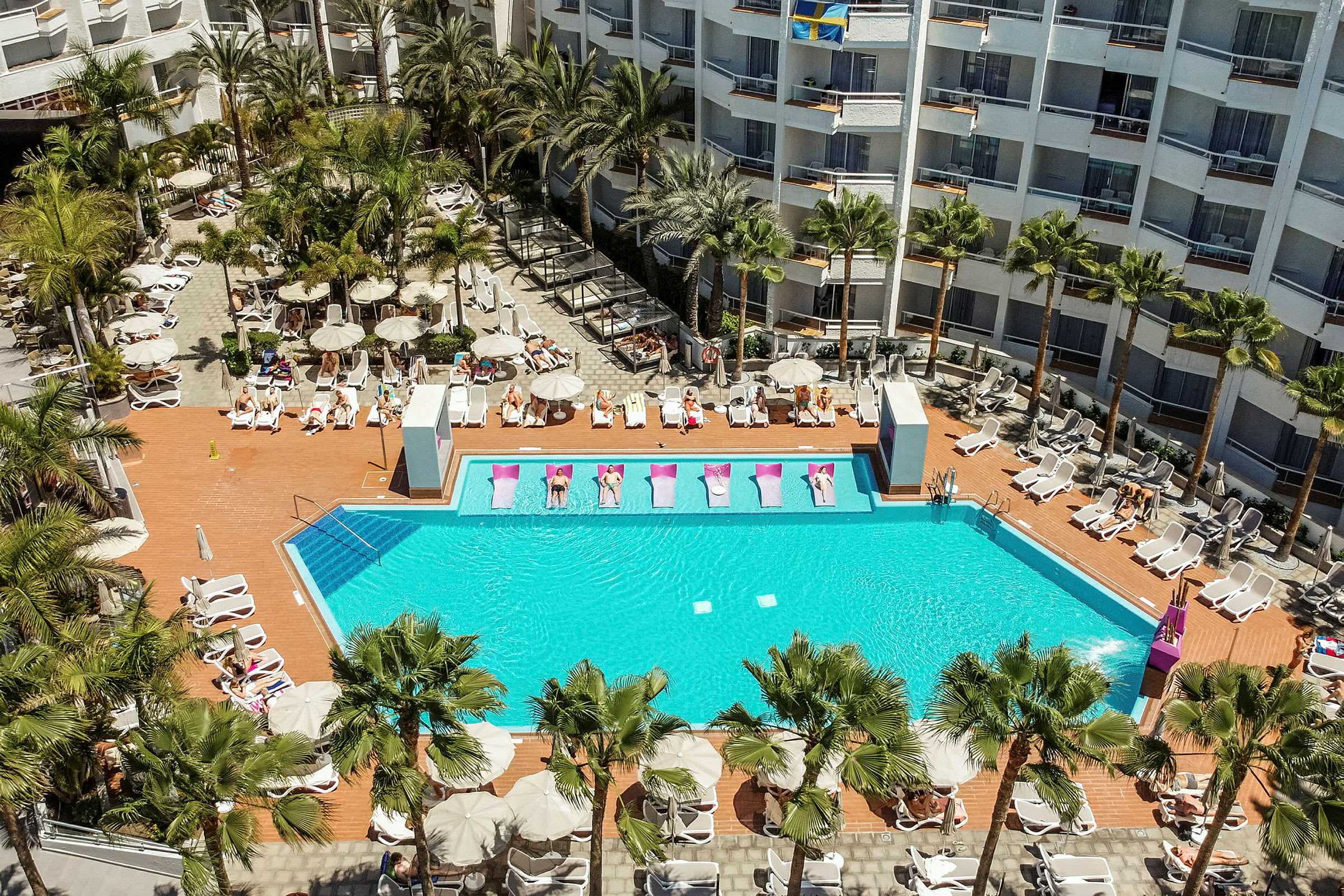 Hotelpool med liggestole og omkringliggende bygninger. (AI-genereret tekst)