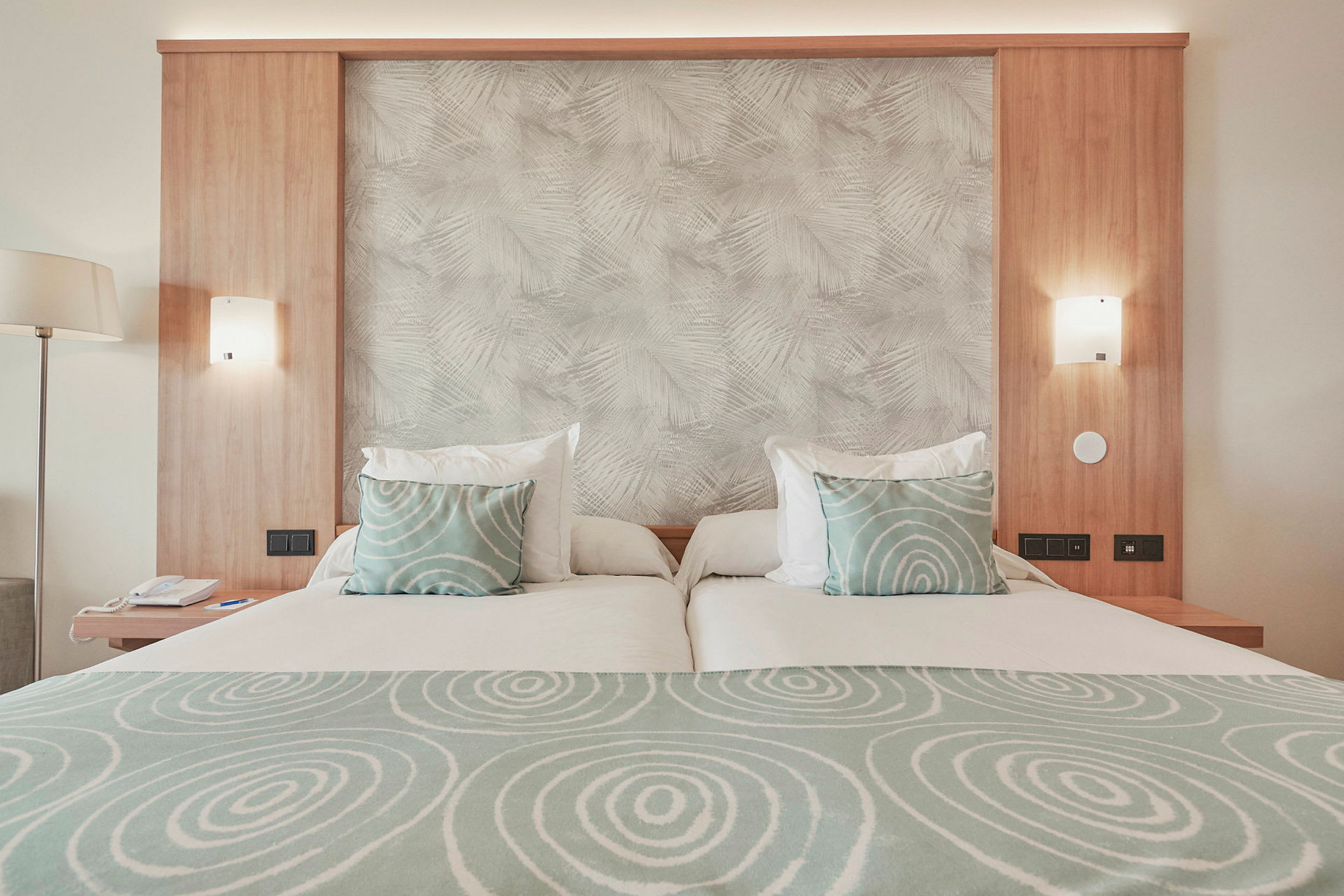 Hotelseng med lys træpanel, grønne puder med cirkelmønster og matchende sengetøj. (AI-genereret tekst)