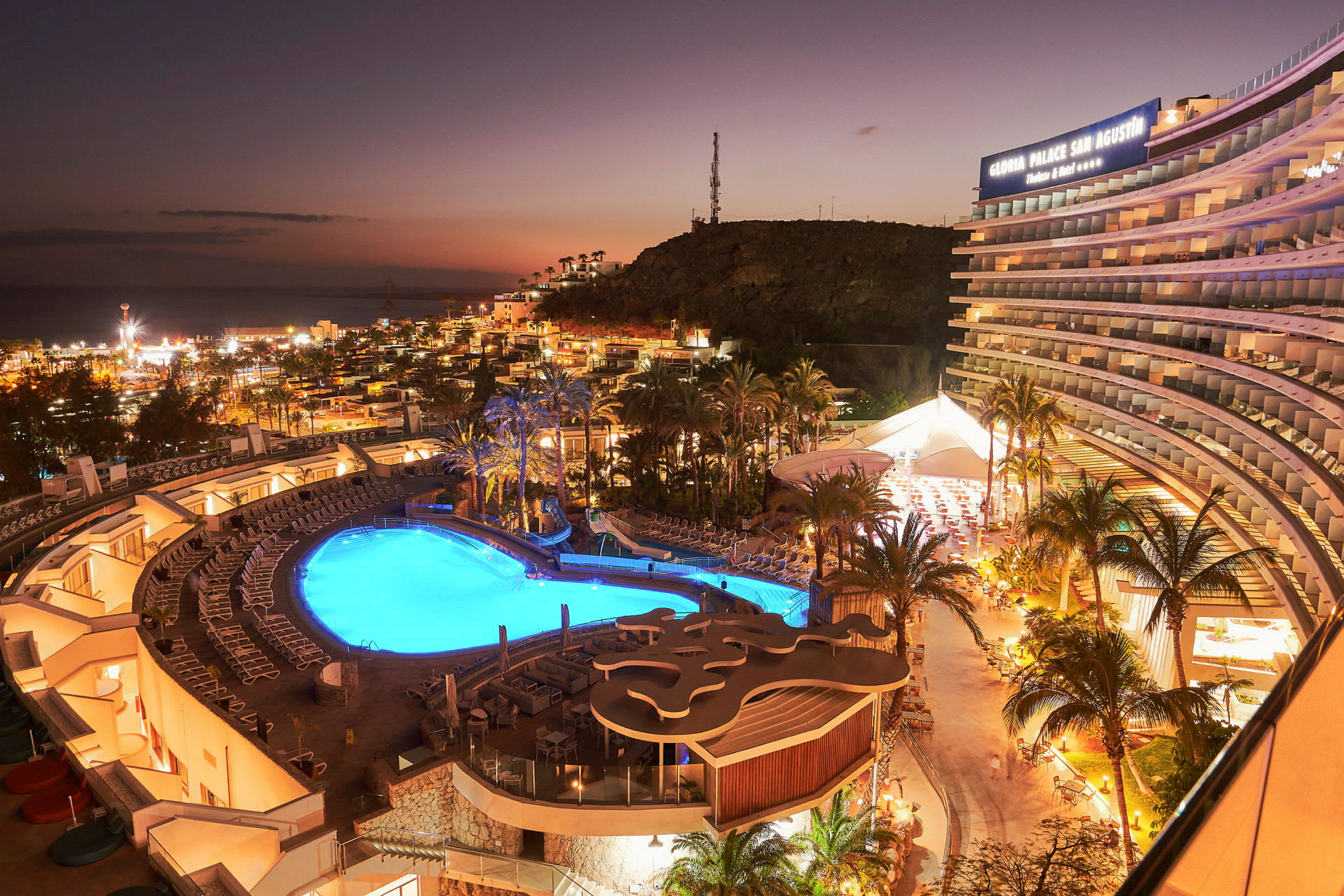 Hotelområde med oplyst pool og fremtrædende bygning ved solnedgang. (AI-genereret tekst)