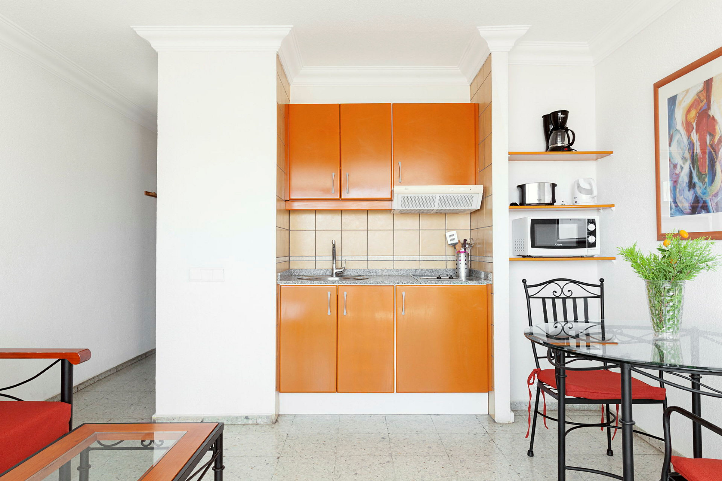 Et køkken med orange skabe, en køkkenvask og husholdningsapparater, ved siden af et spiseområde med glasbord og metalstole. (AI-genereret tekst)