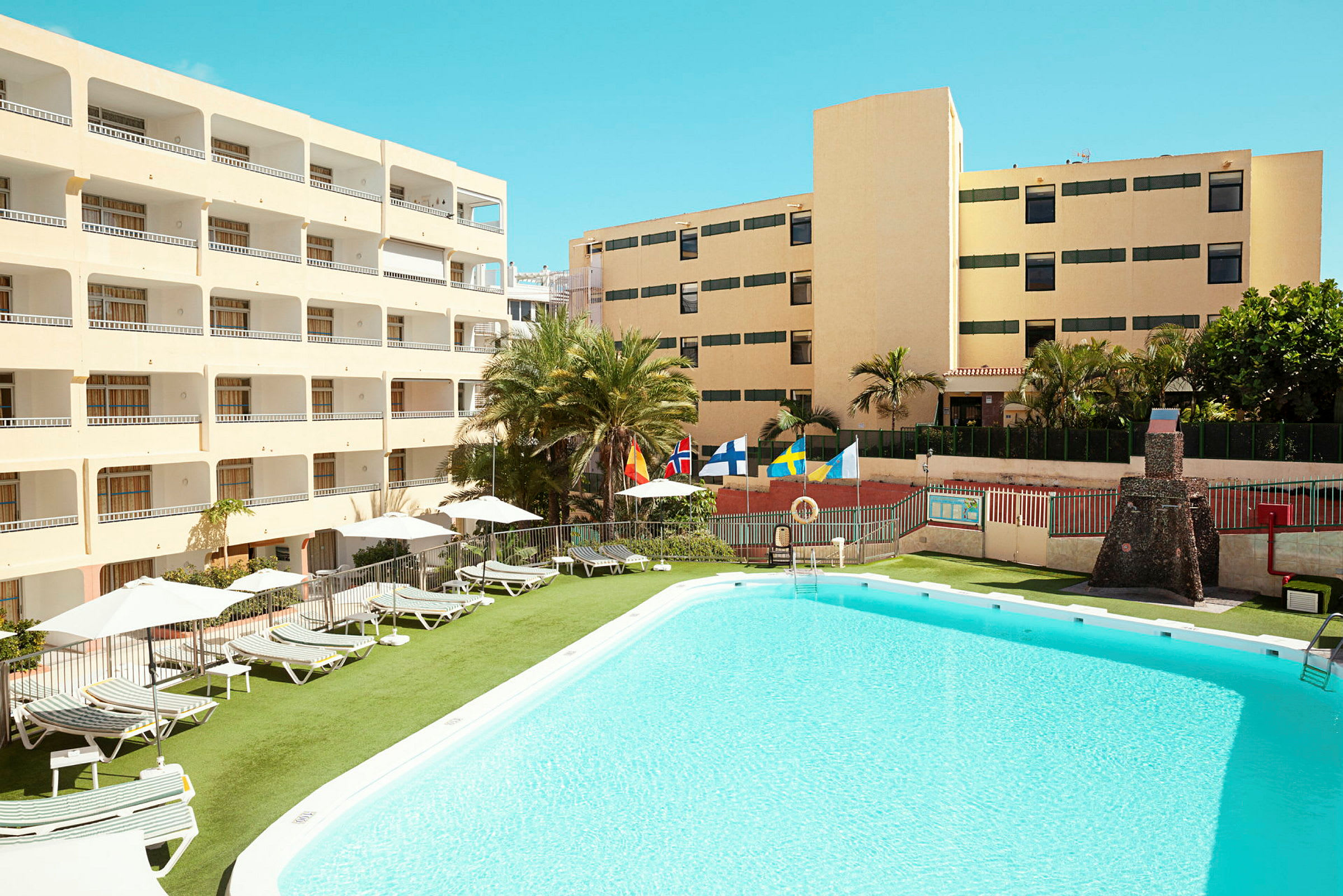 Et billede af et hotelområde med pool og liggestole. Bygninger omkranser området. (AI-genereret tekst)