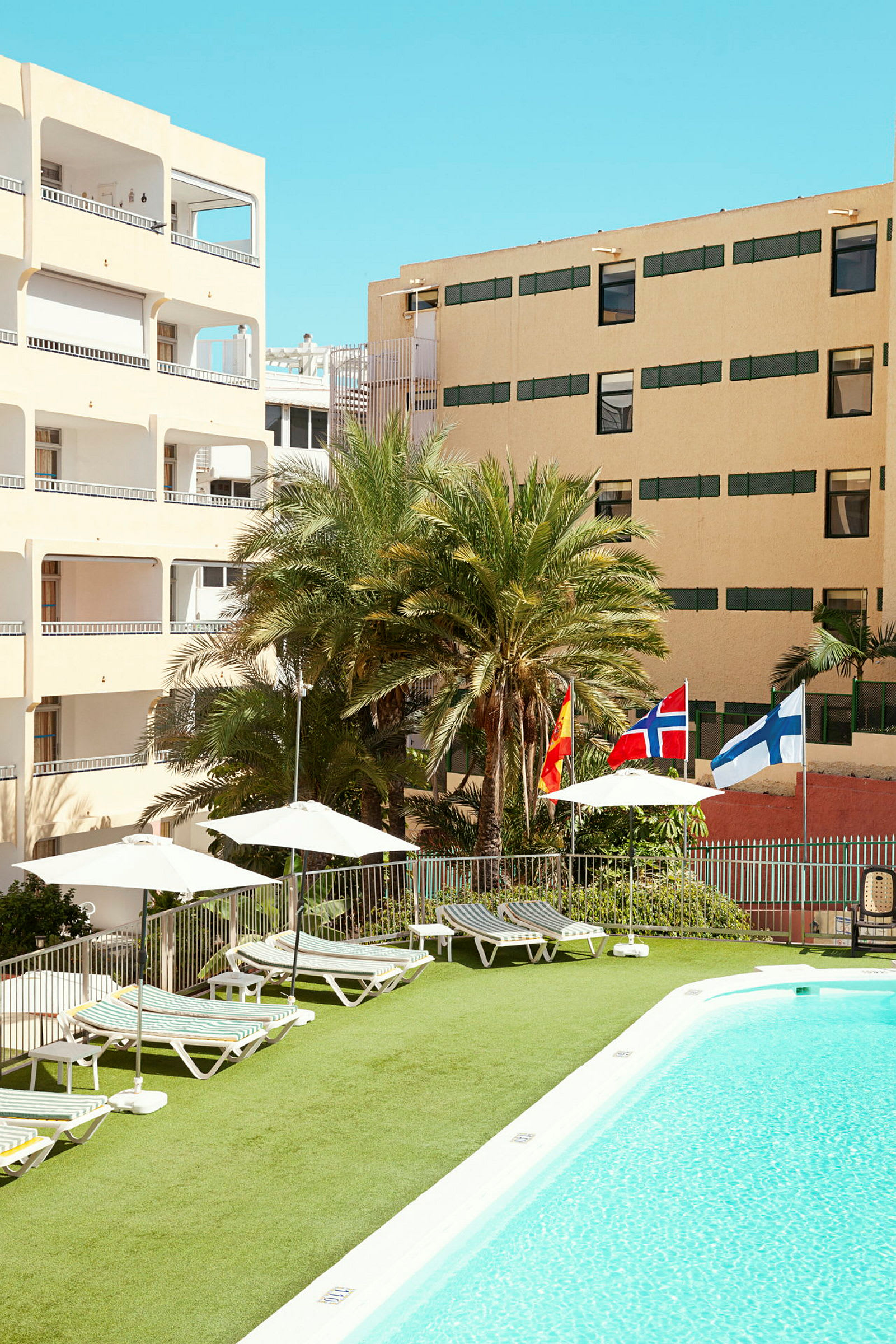 Billede af en hotelpool med liggestole, parasoller og flag fra Spanien, Norge og Finland. Baggrunden viser hotelbygninger og palmer. (AI-genereret tekst)