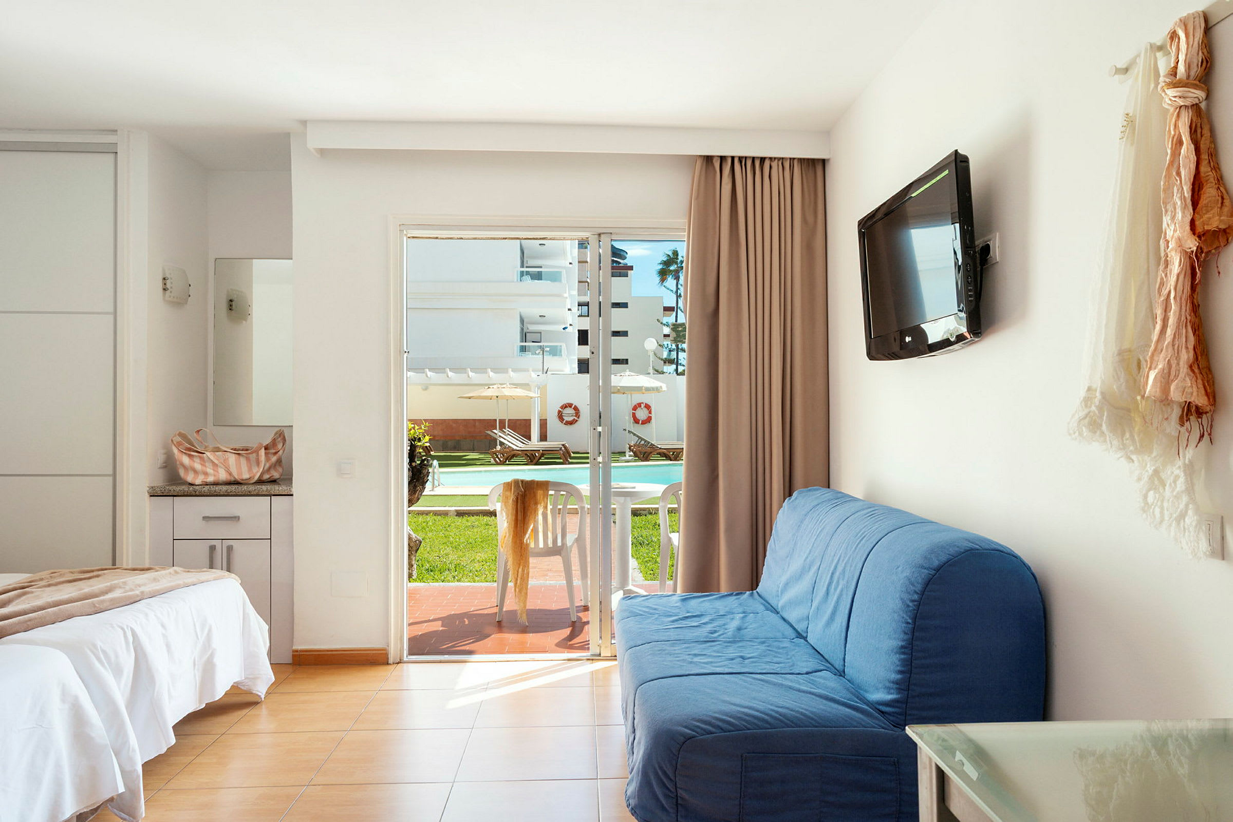Et hotelværelse med en opredt seng, blå sofa, vægmonteret TV og en glasskydedør, der fører ud til en terrasse med bord og stole. Udsigten viser en udendørspool og liggestole. (AI-genereret tekst)