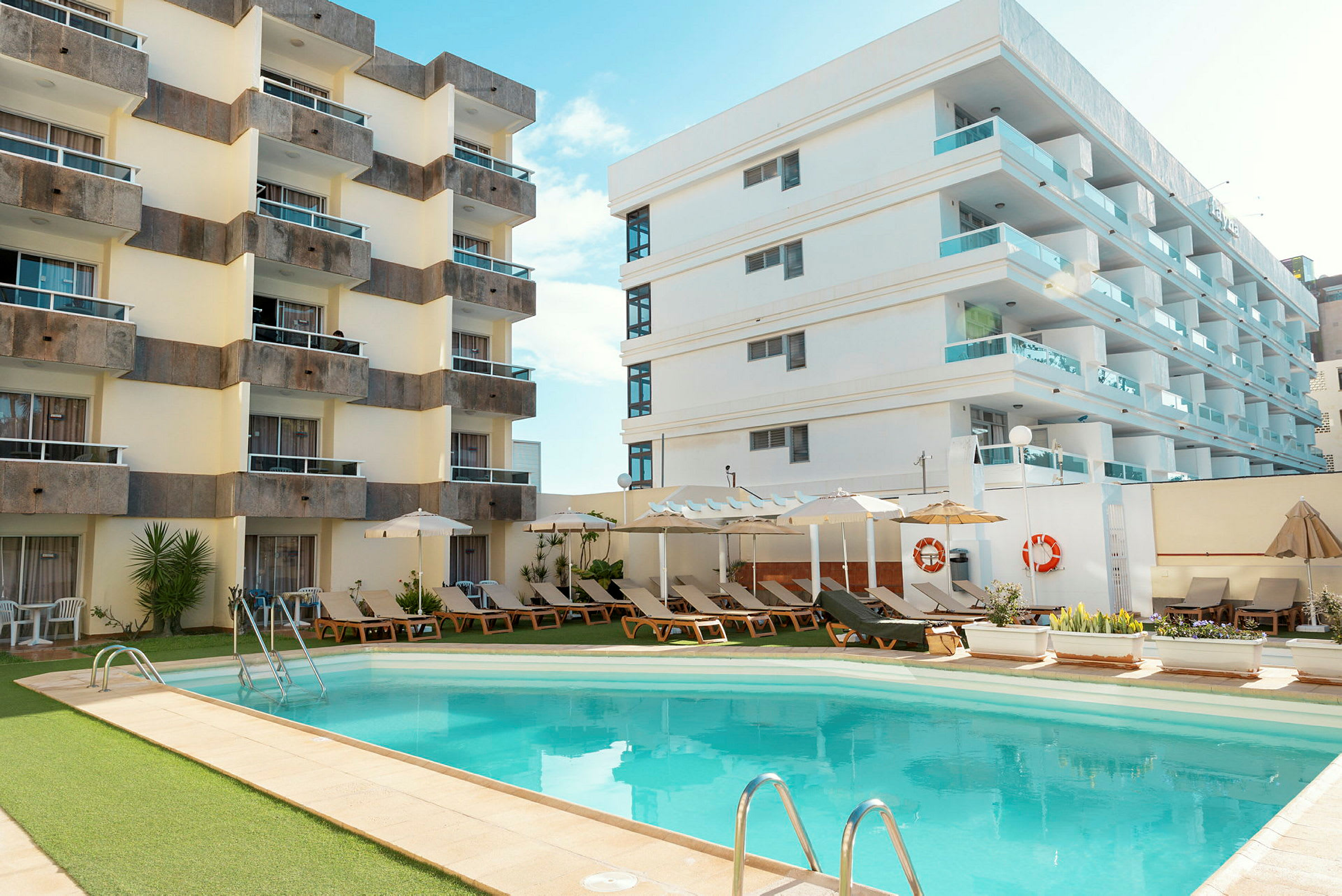 Hotelbygninger ved en udendørspool omgivet af liggestole og parasoller. (AI-genereret tekst)