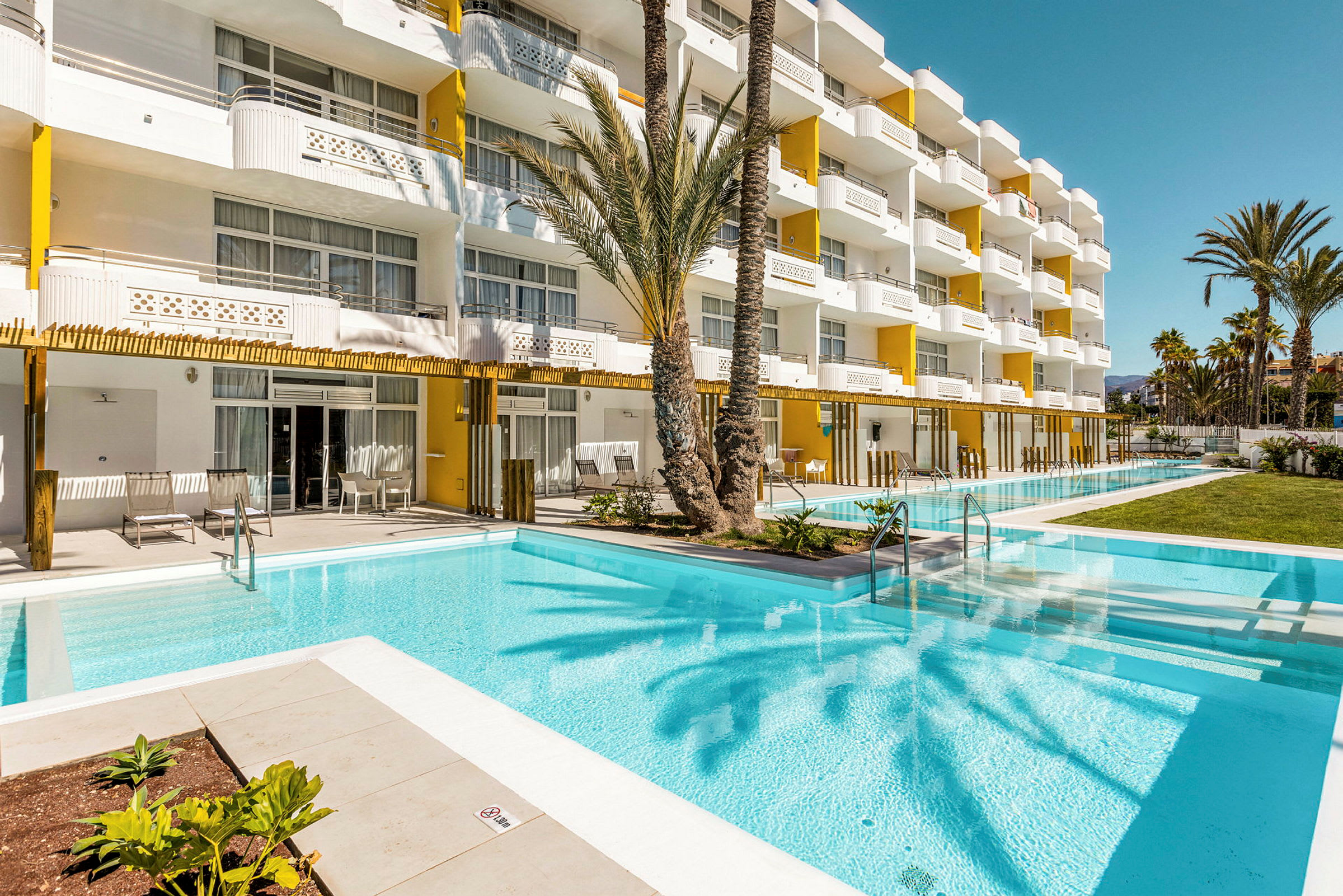 Solbelyst hotel med gul og hvid facade, pool og grønne områder. (AI-genereret tekst)