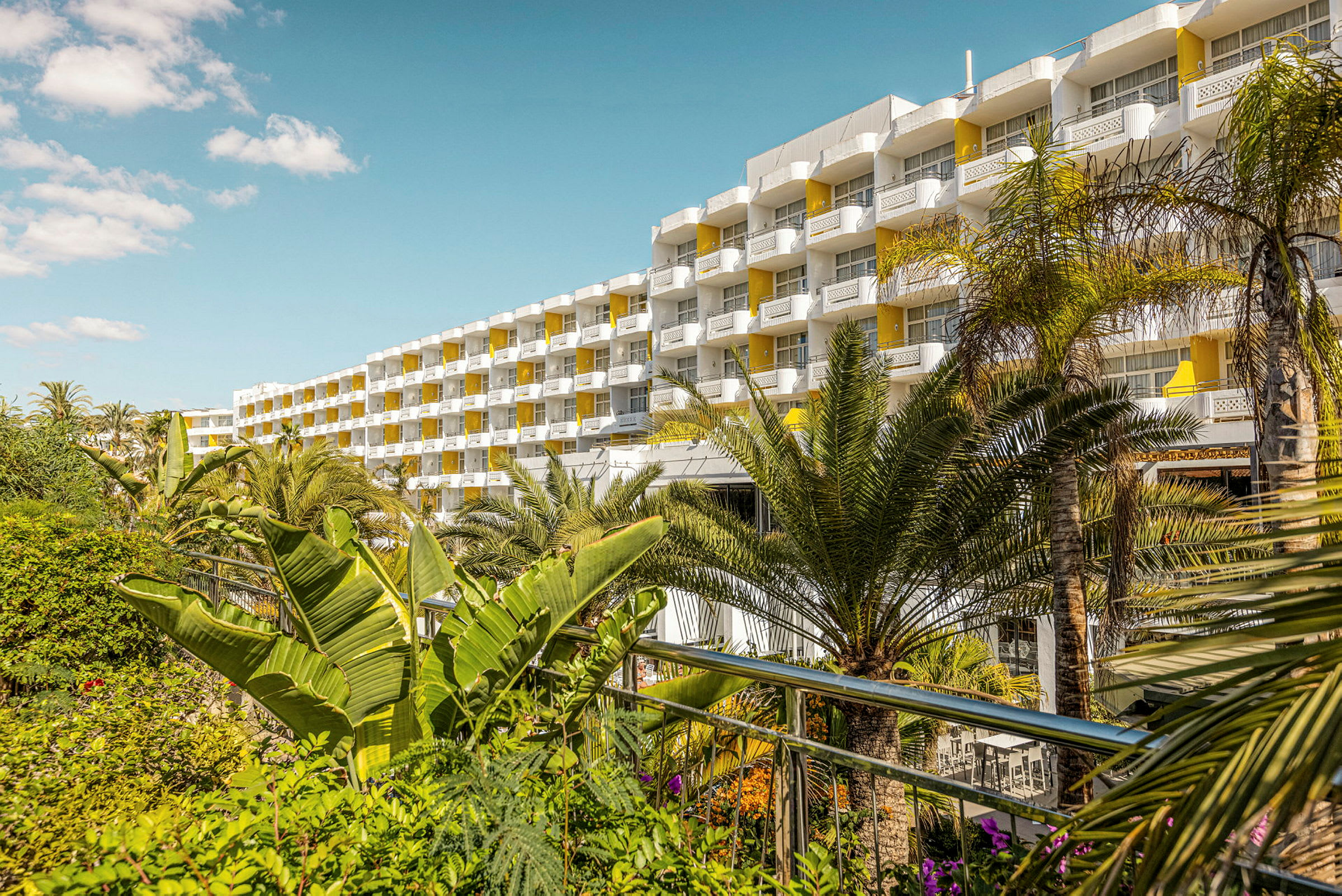 Flereårigt hotel med gule altaner omgivet af frodige palmer. (AI-genereret tekst)