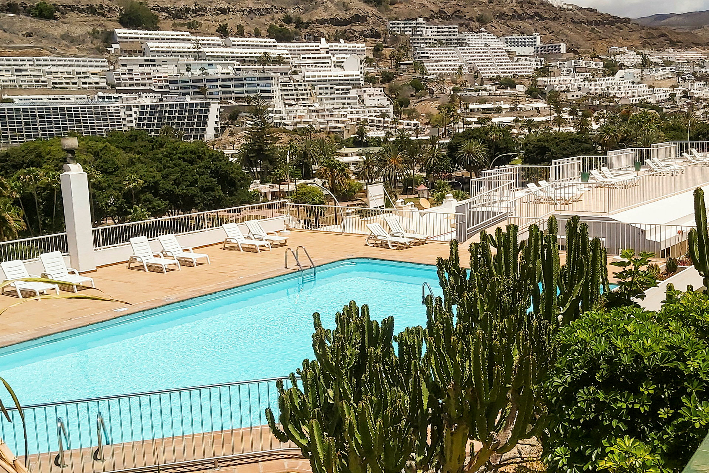 Udendørs swimmingpool med liggestole og grønt, omgivet af bygninger og bjerge. (AI-genereret tekst)