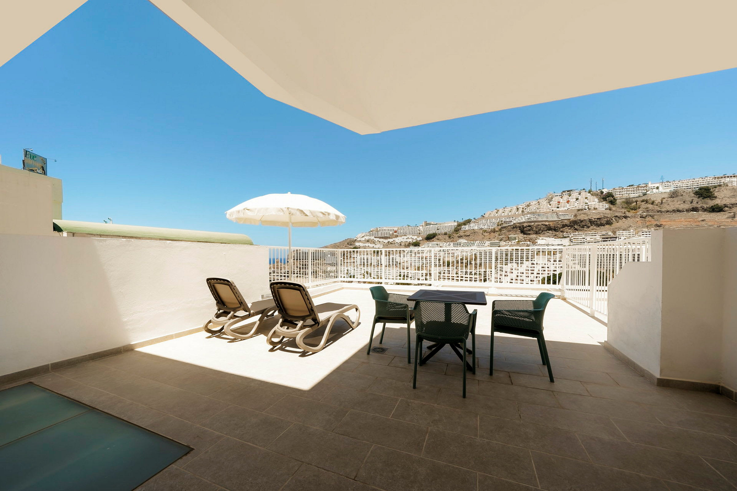En terrasse med liggestole, parasol og havemøbler med udsigt mod solen og en bjergskråning. (AI-genereret tekst)