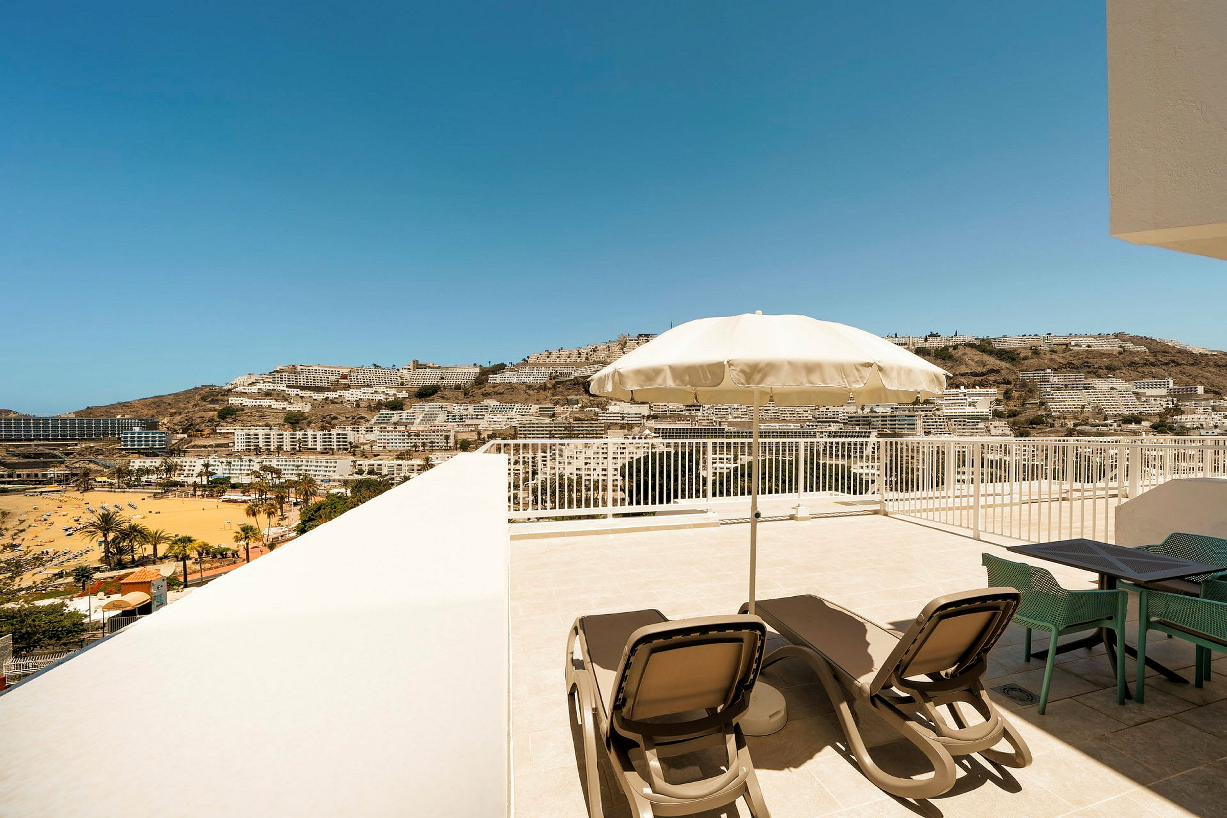 Terrasse med solsenge og parasol, udsigt over en strand og by mod bjerge i det fjerne. (AI-genereret tekst)