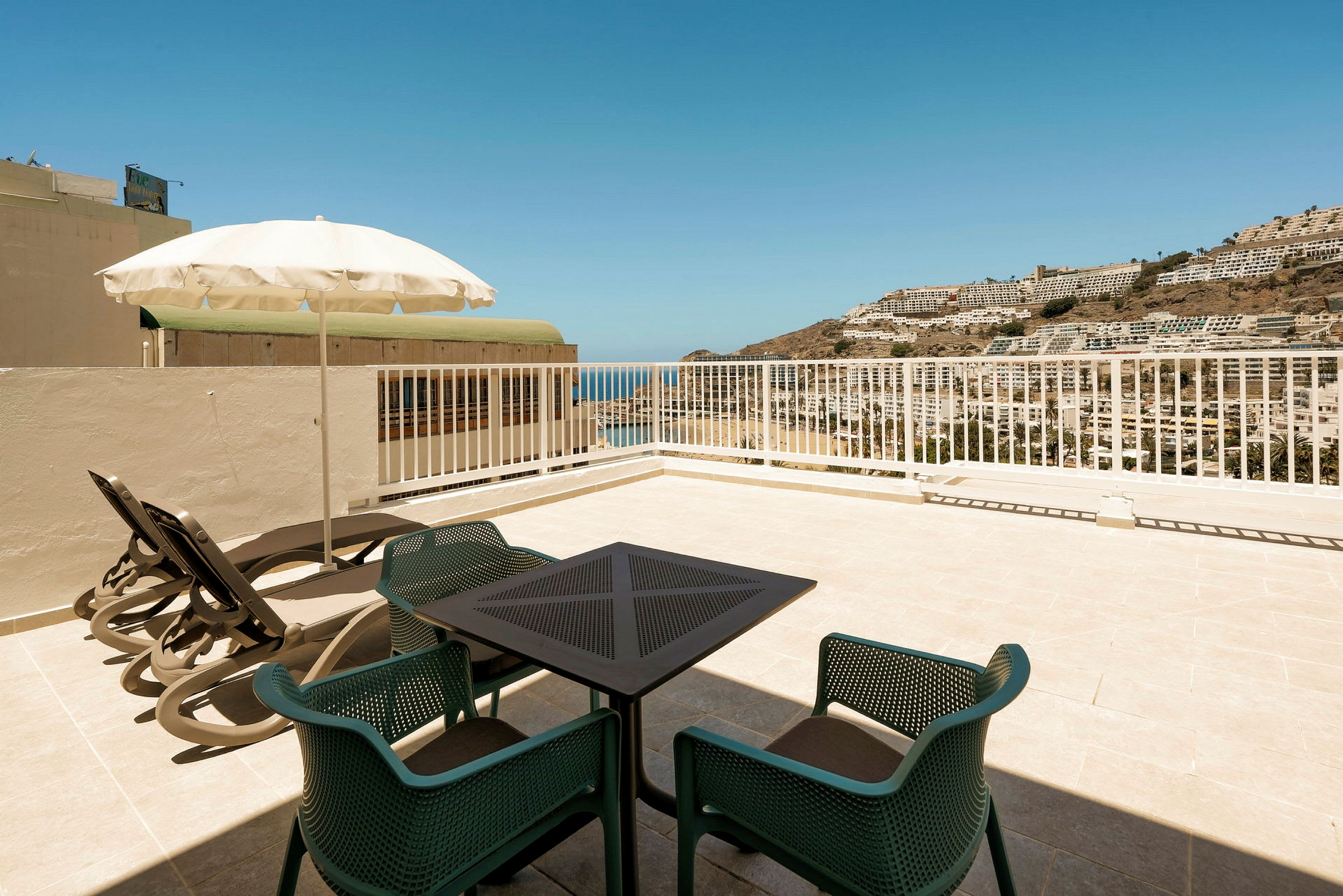 Udsigt fra en terrasse med bord, stole og liggestole mod havet og bygninger på en bjergskråning. (AI-genereret tekst)