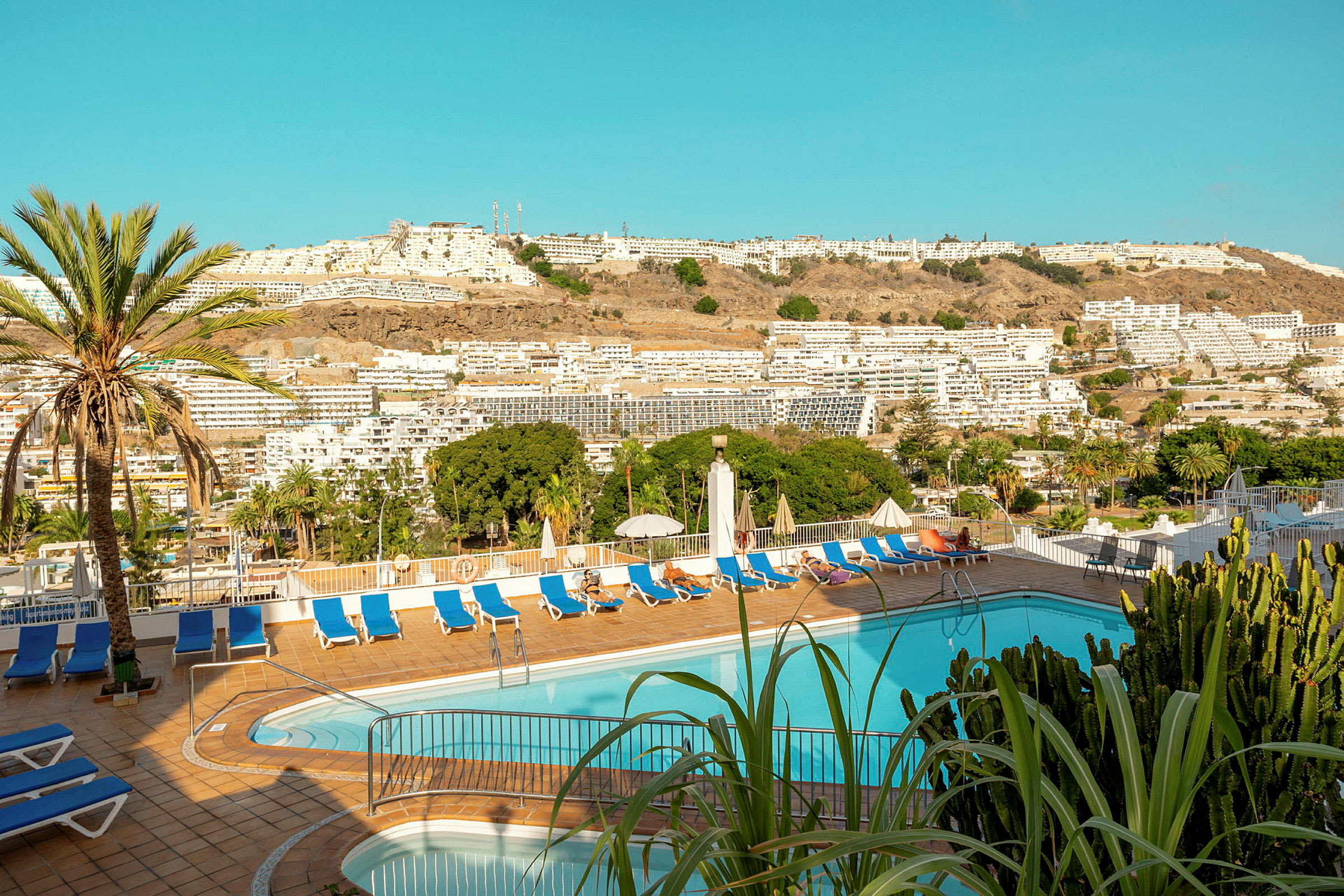 Billede af en hotelpool med liggestole og parasoller. I baggrunden ses bygninger og grønne områder. (AI-genereret tekst)