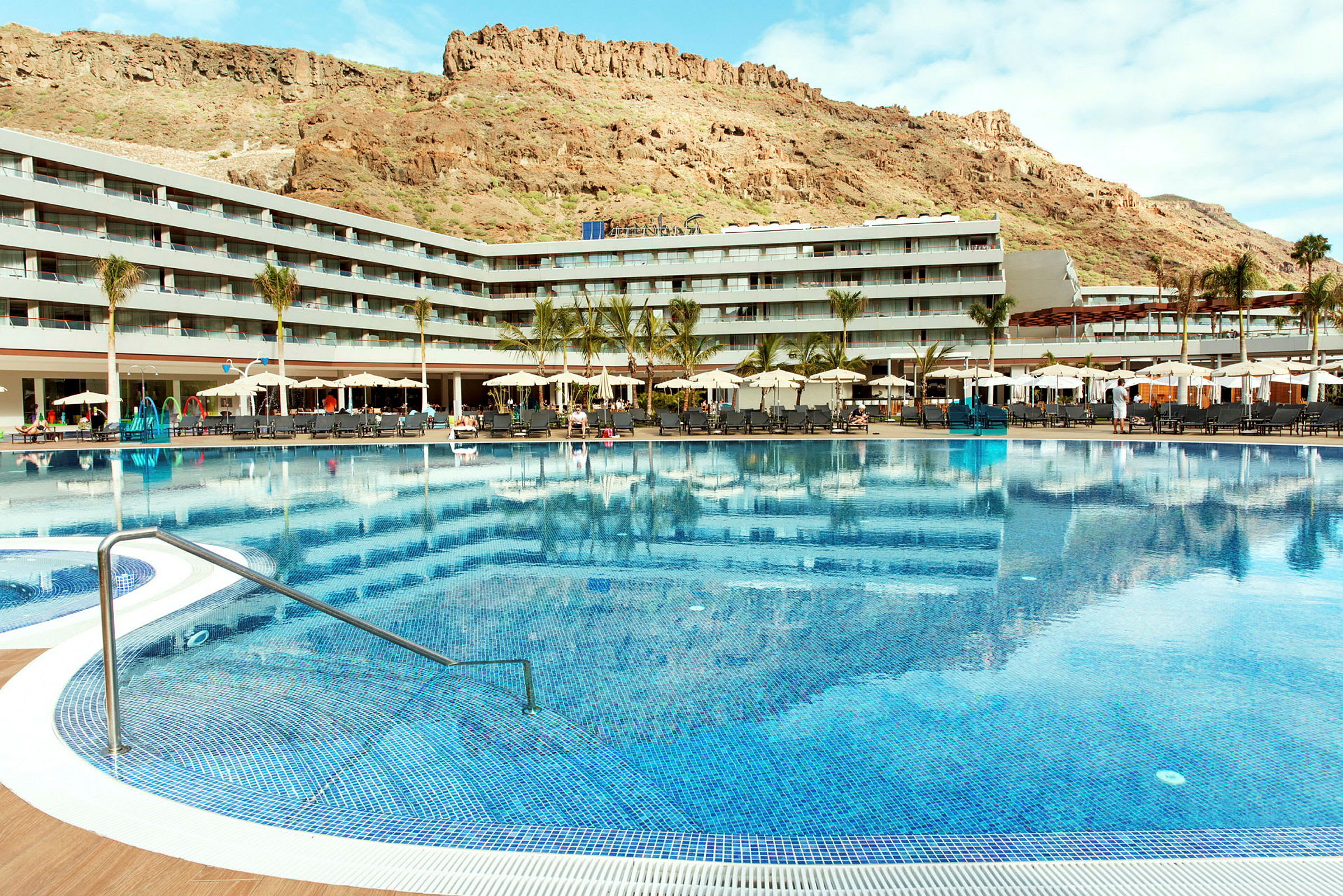 Stor udendørs pool foran hotelbygning med bjerge i baggrunden. (AI-genereret tekst)