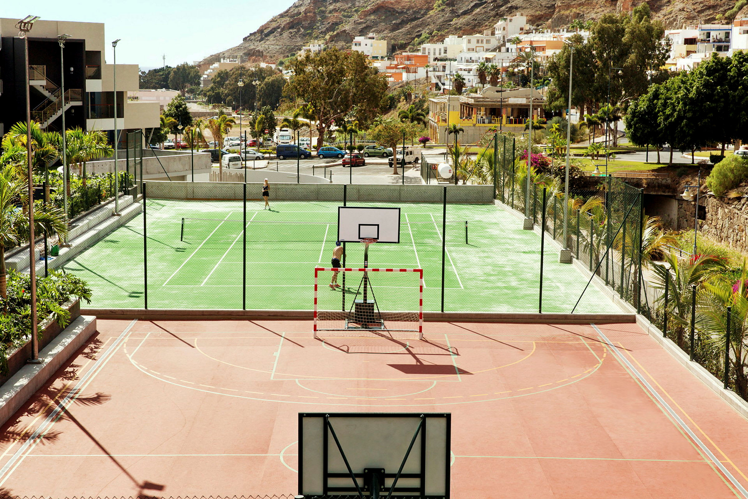 Et billede af en farverig sportsplads med basketballmål og tennisbaner, omgivet af grønne områder, bygninger og bakker. (AI-genereret tekst)