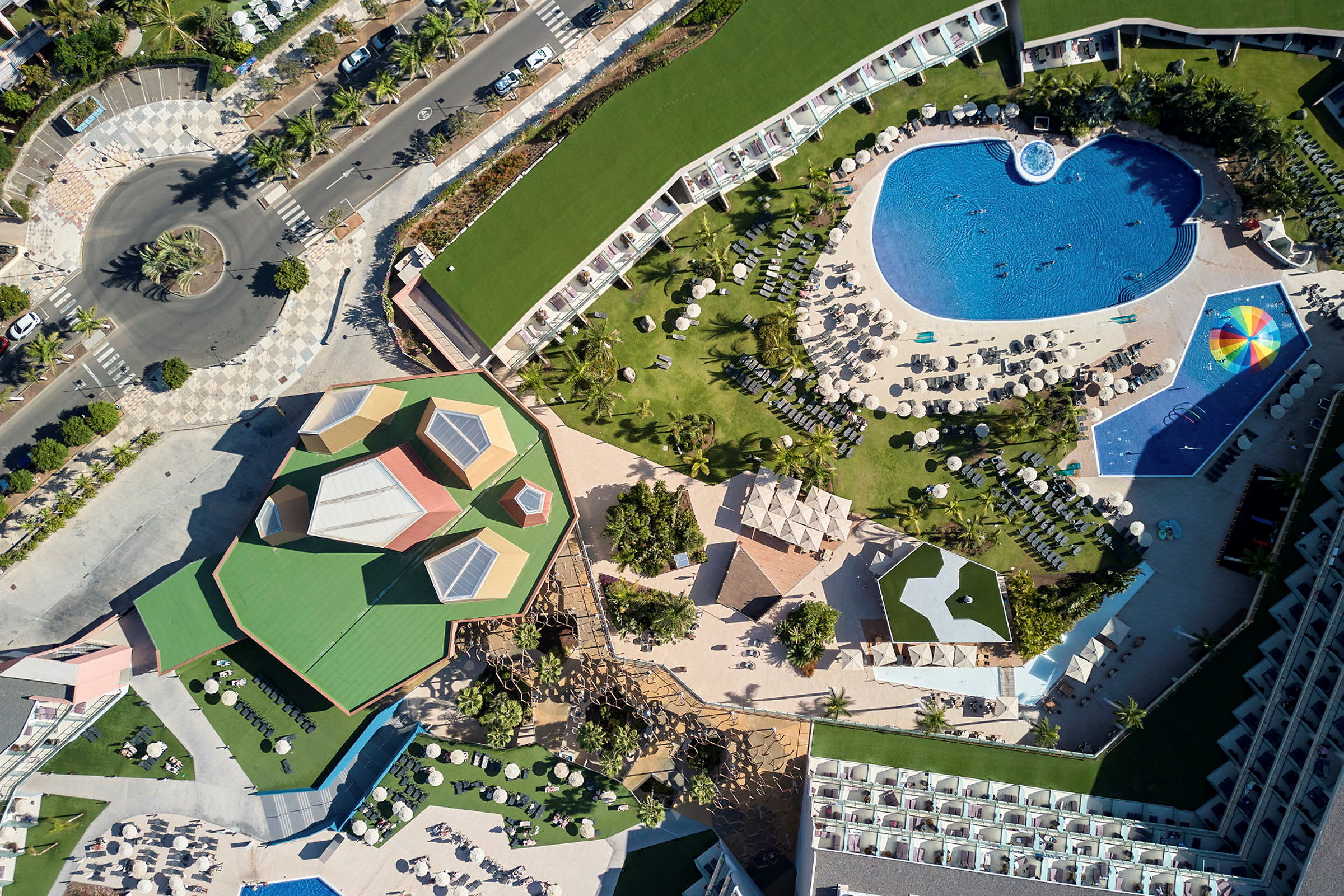 Luftfoto af en ferieresort med poolområde, grønne områder og bygninger. (AI-genereret tekst)