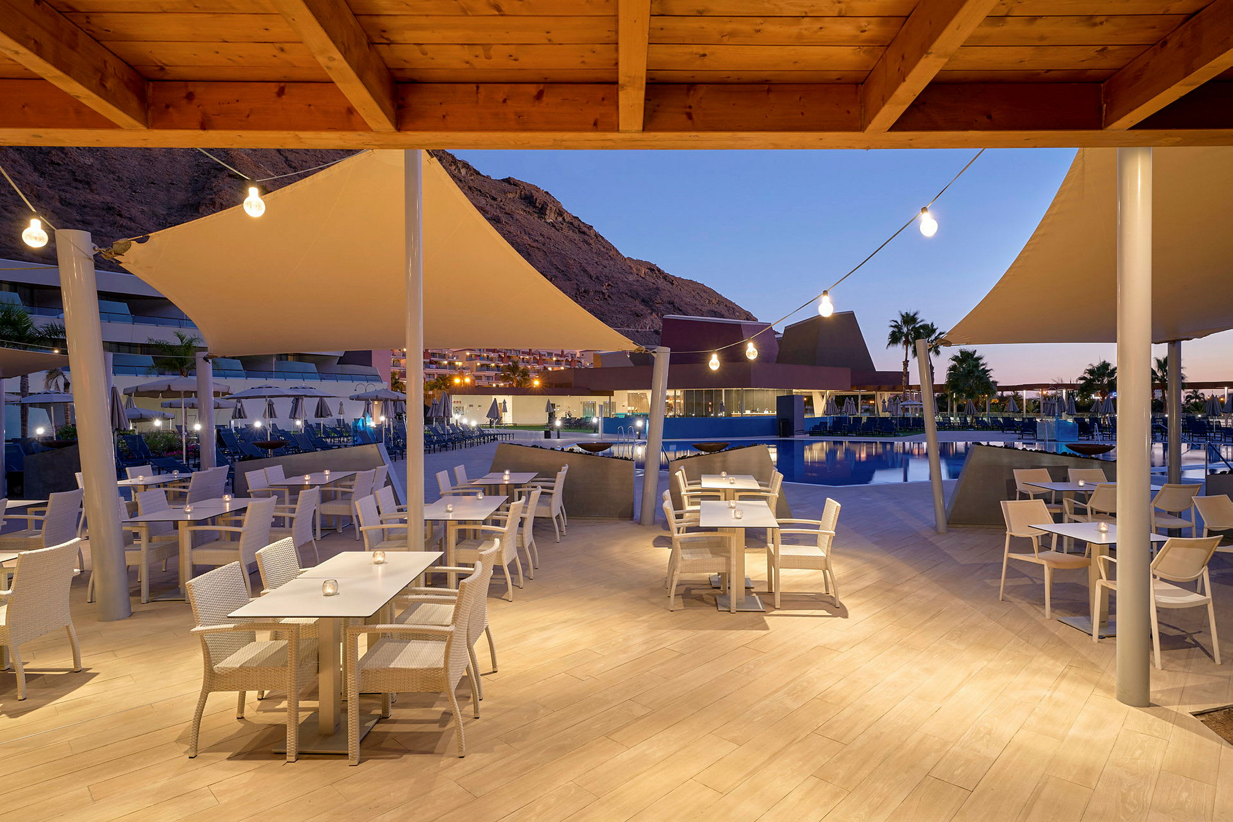 Udendørs restaurant med hvide stole og borde ved en pool, aftenbelysning og bjerge i baggrunden. (AI-genereret tekst)