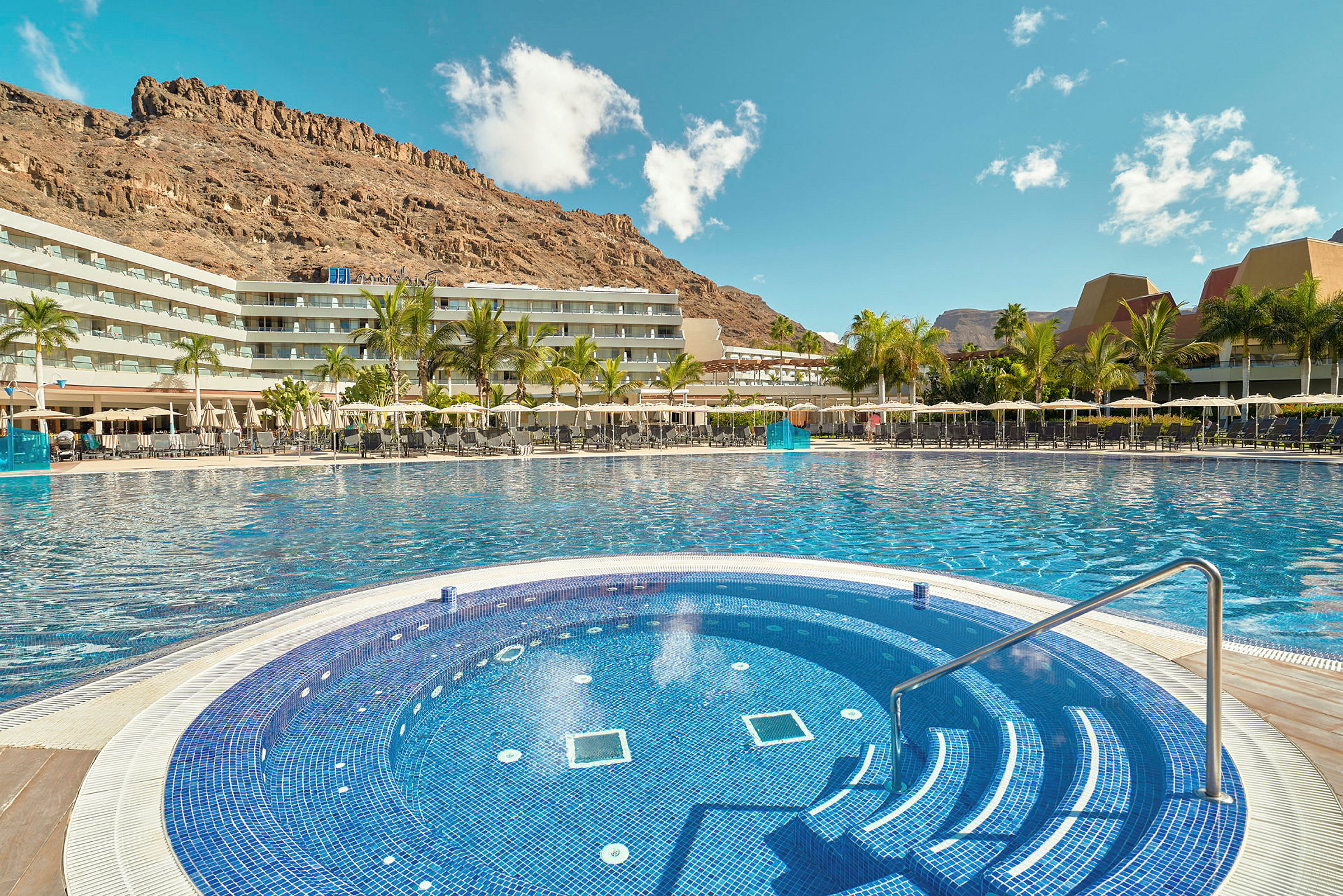 Hotellets poolområde med en mindre rund pool i forgrunden og omkringliggende bygninger samt bjerge i baggrunden. (AI-genereret tekst)