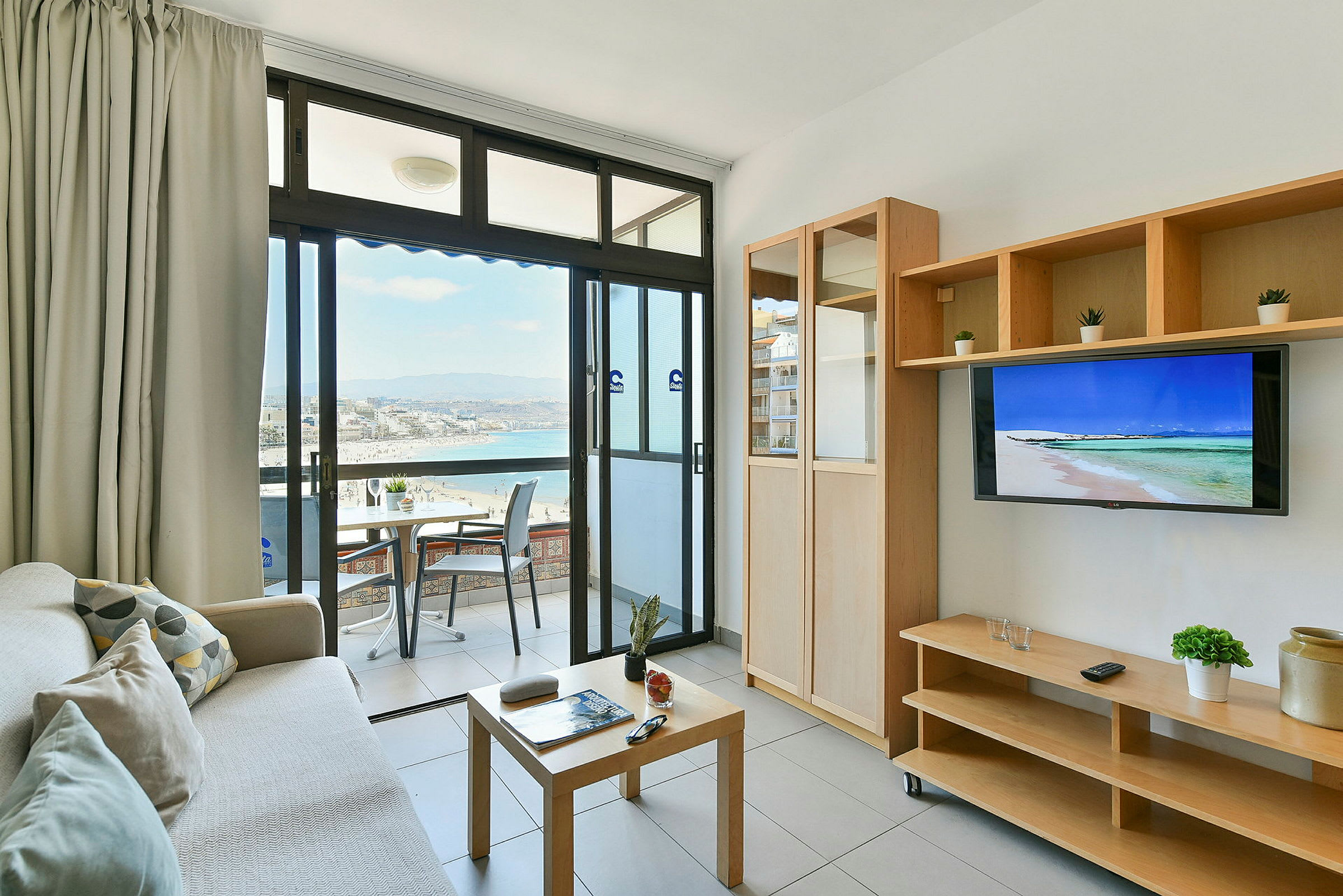 En stue med adgang til en balkon med udsigt over en strand og havet. Rummet har møbler som en sofa, et sofabord og et TV. (AI-genereret tekst)