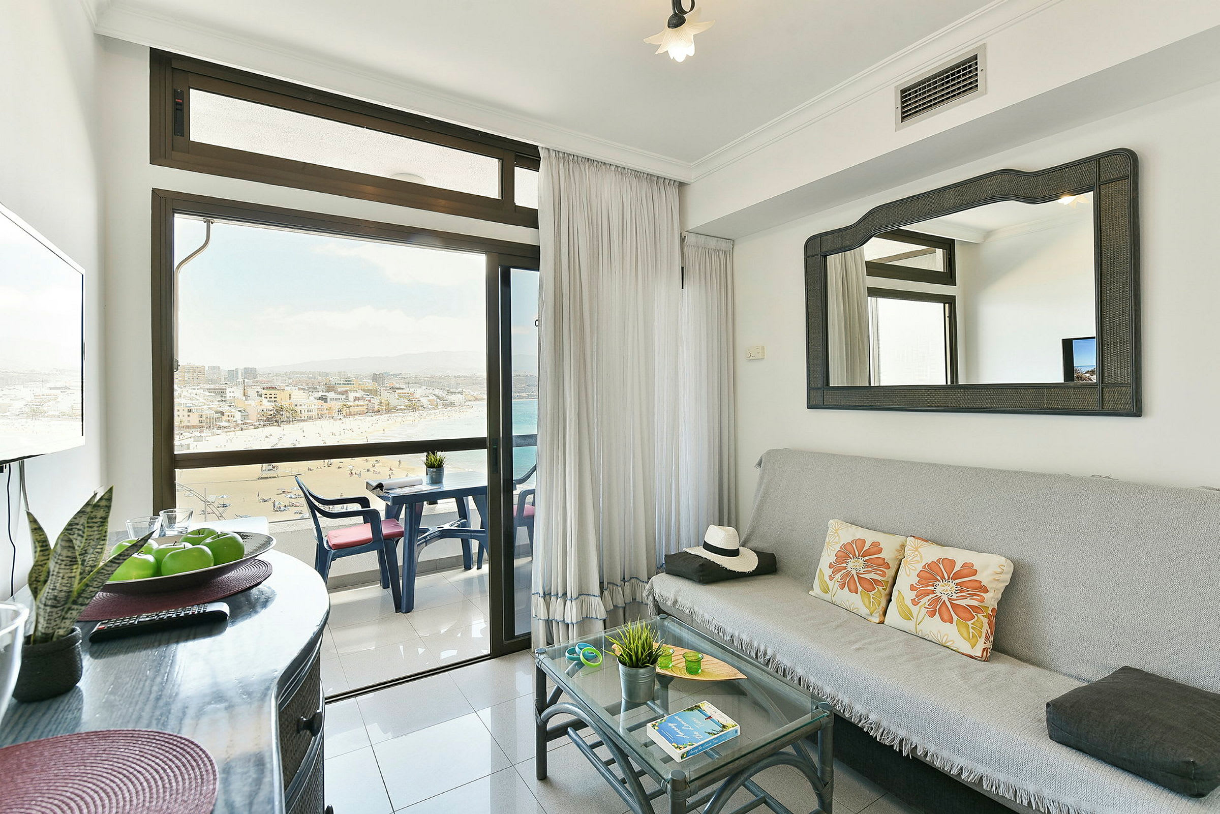 Stue med glasbord, sofa, spejl og altan med udsigt til stranden. (AI-genereret tekst)