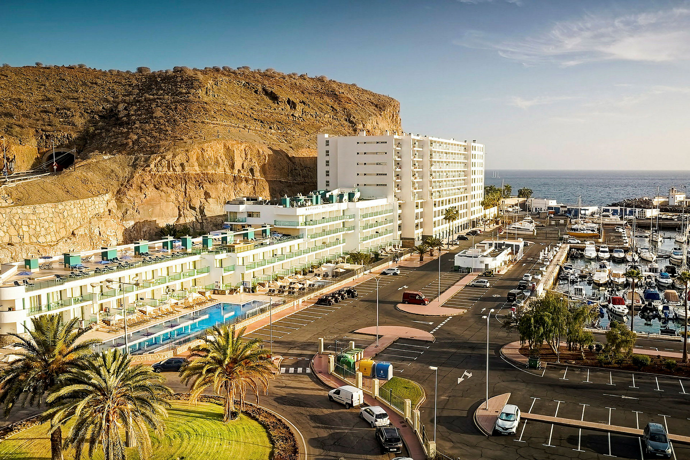 Hotel ved klippekant med parkeringsplads og båd havn ved havet. (AI-genereret tekst)