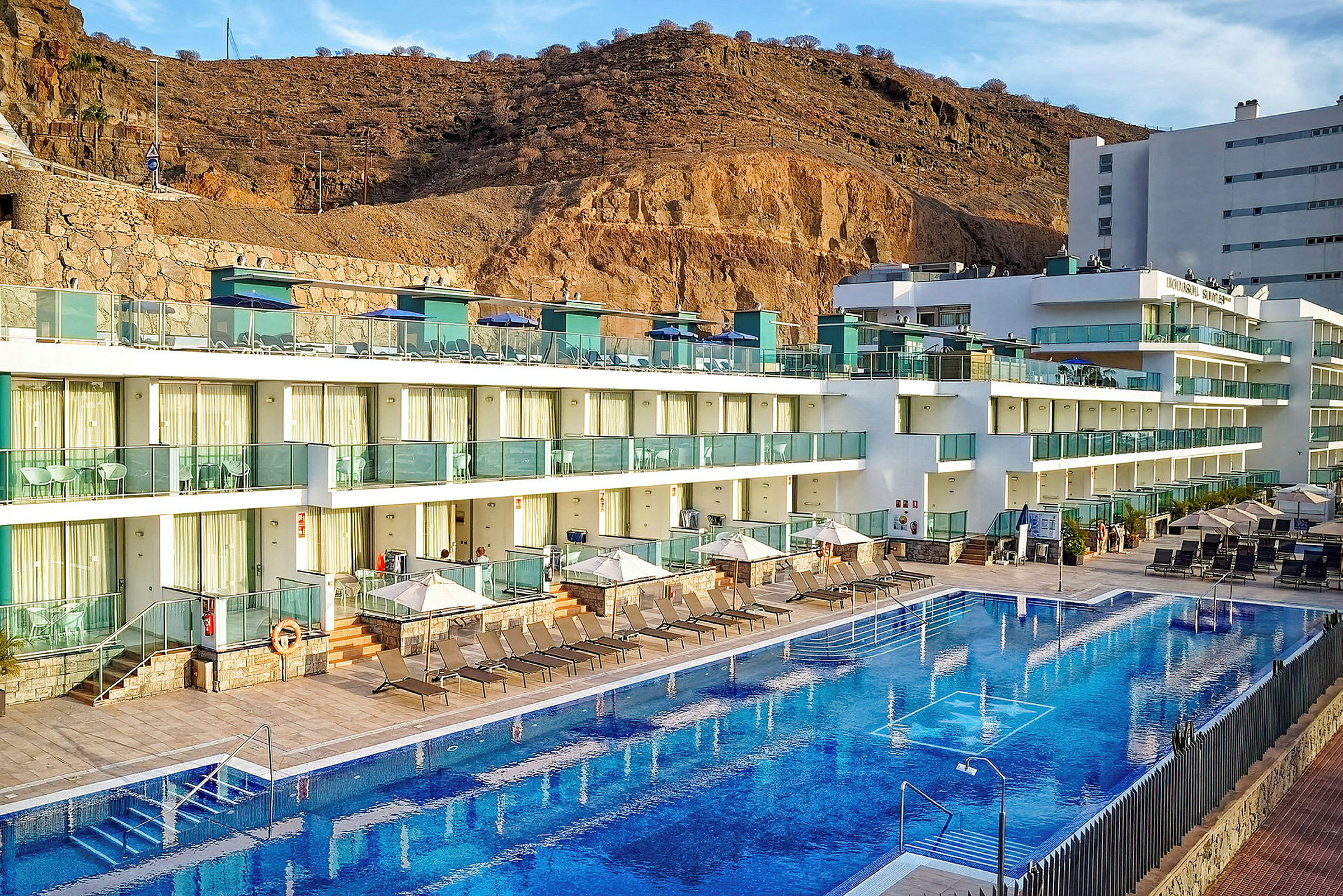 Hotelkompleks med flere etager og glasfacader, liggestole og parasoller ved en udendørs pool, omgivet af bjerge. (AI-genereret tekst)