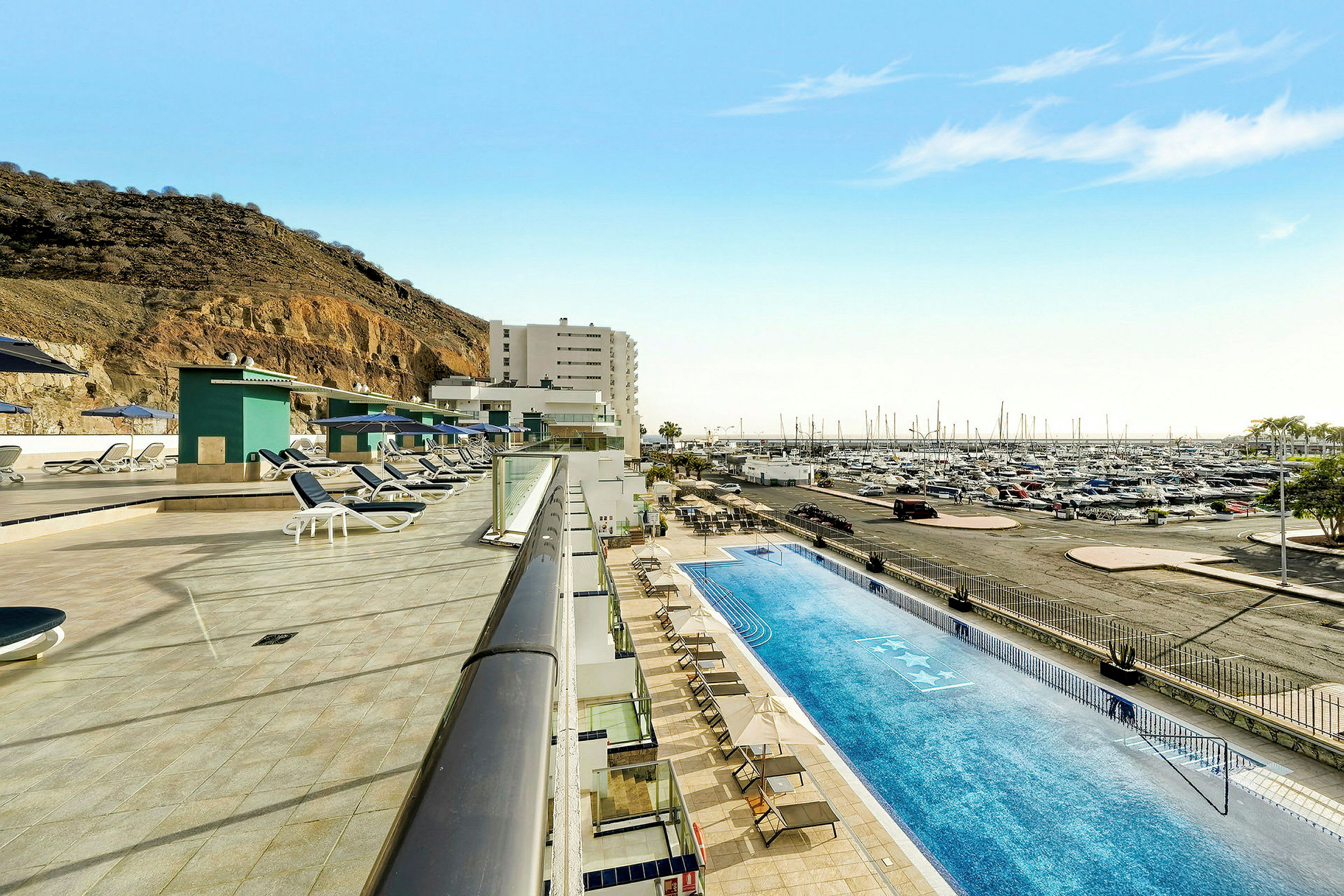 Poolområde med liggestole ved siden af en marina og en bjergskråning. (AI-genereret tekst)