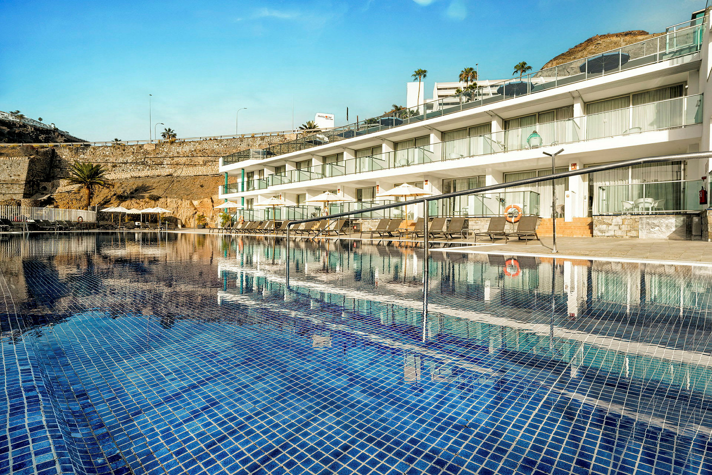 En swimmingpool med blå fliser foran en række hvide hotelværelser. (AI-genereret tekst)