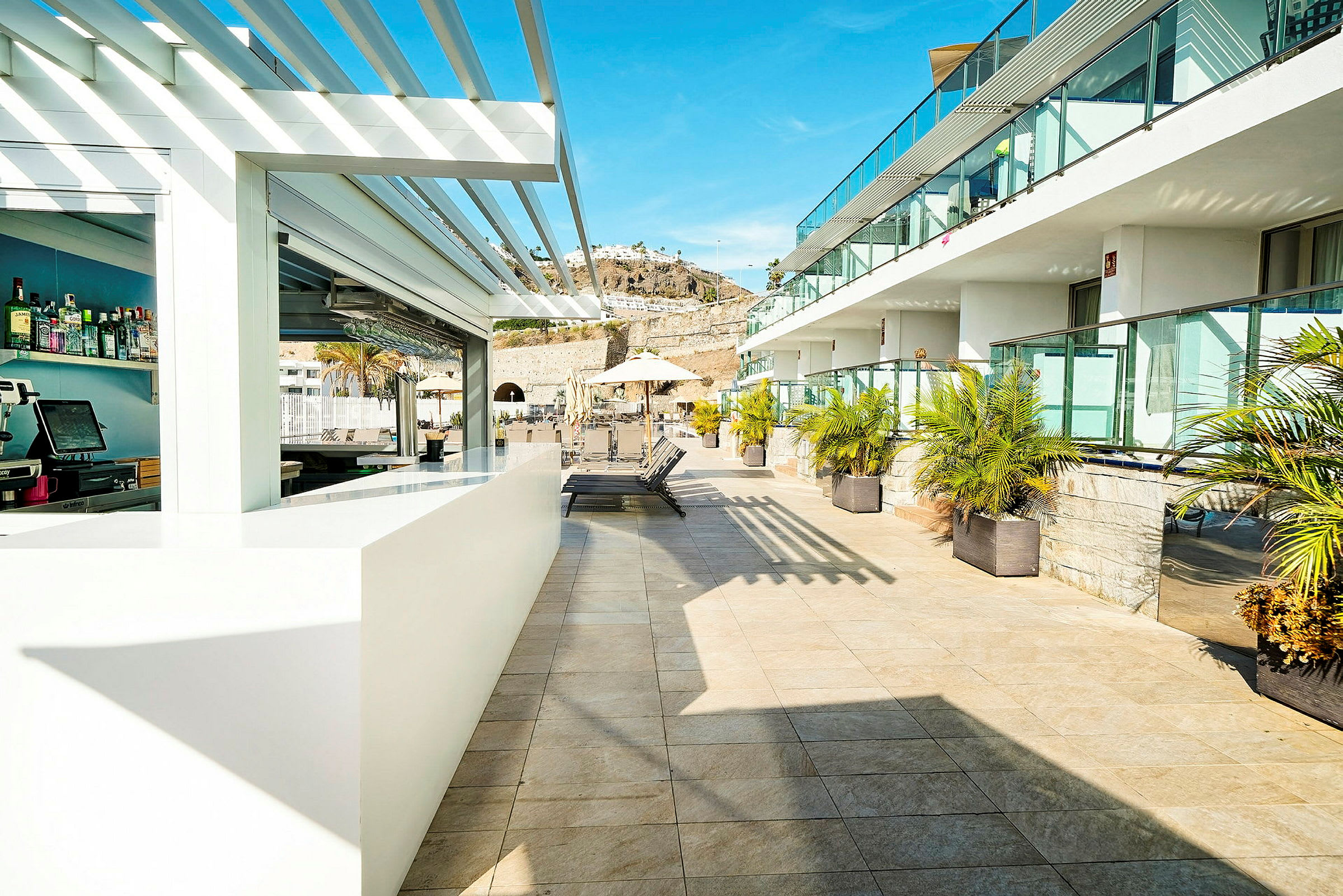 Billede af en solrig hotelterrasse med et barområde og liggestole, omgivet af palmer og glasrækværk. (AI-genereret tekst)