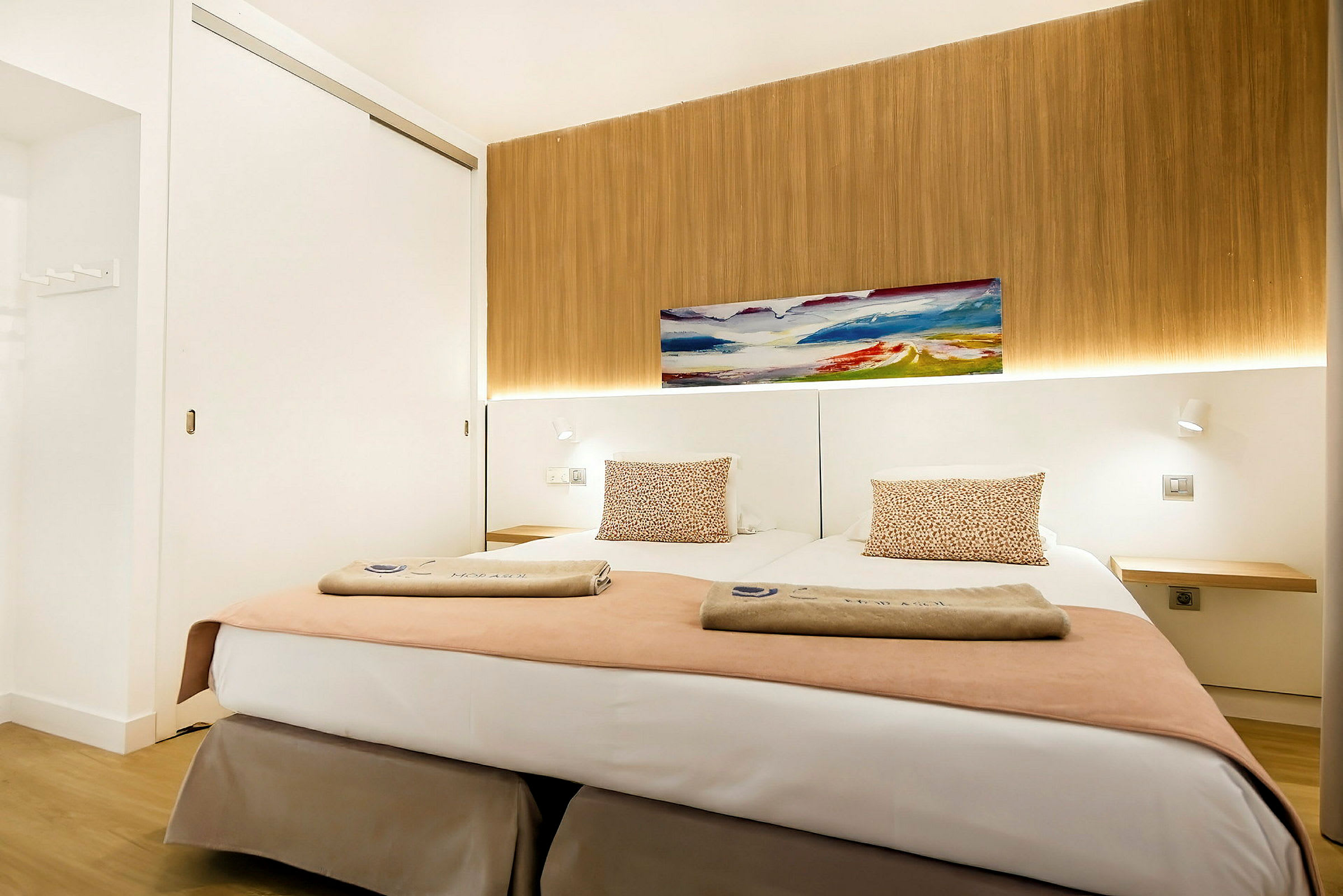 Et moderne hotelværelse med to enkeltsenge, dekoreret med puder og håndklæder, et maleri på væggen og en baggrund med træpanel. (AI-genereret tekst)