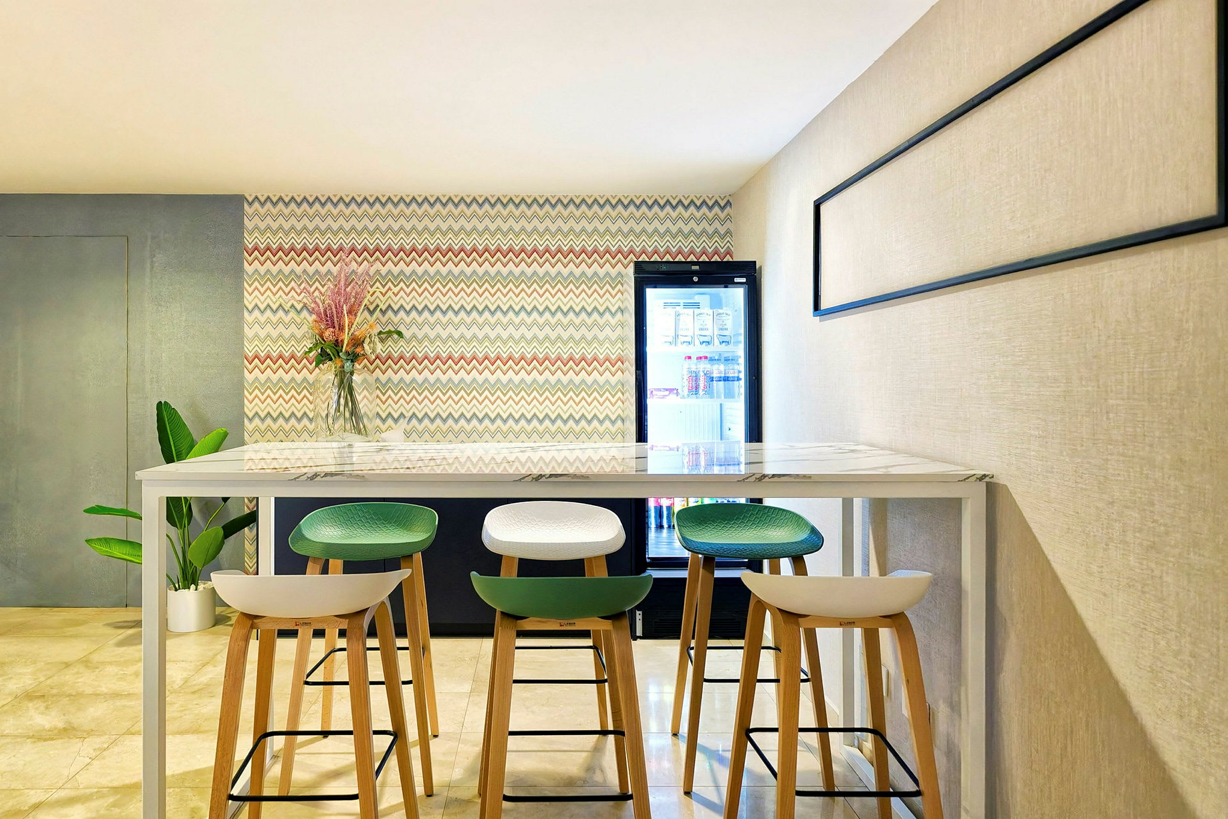 Et moderne rum med høje stole placeret omkring et langbord. På væggen bagved er der et farverigt mønster og en glasvase med blomster. Til højre er der et køleskab. (AI-genereret tekst)