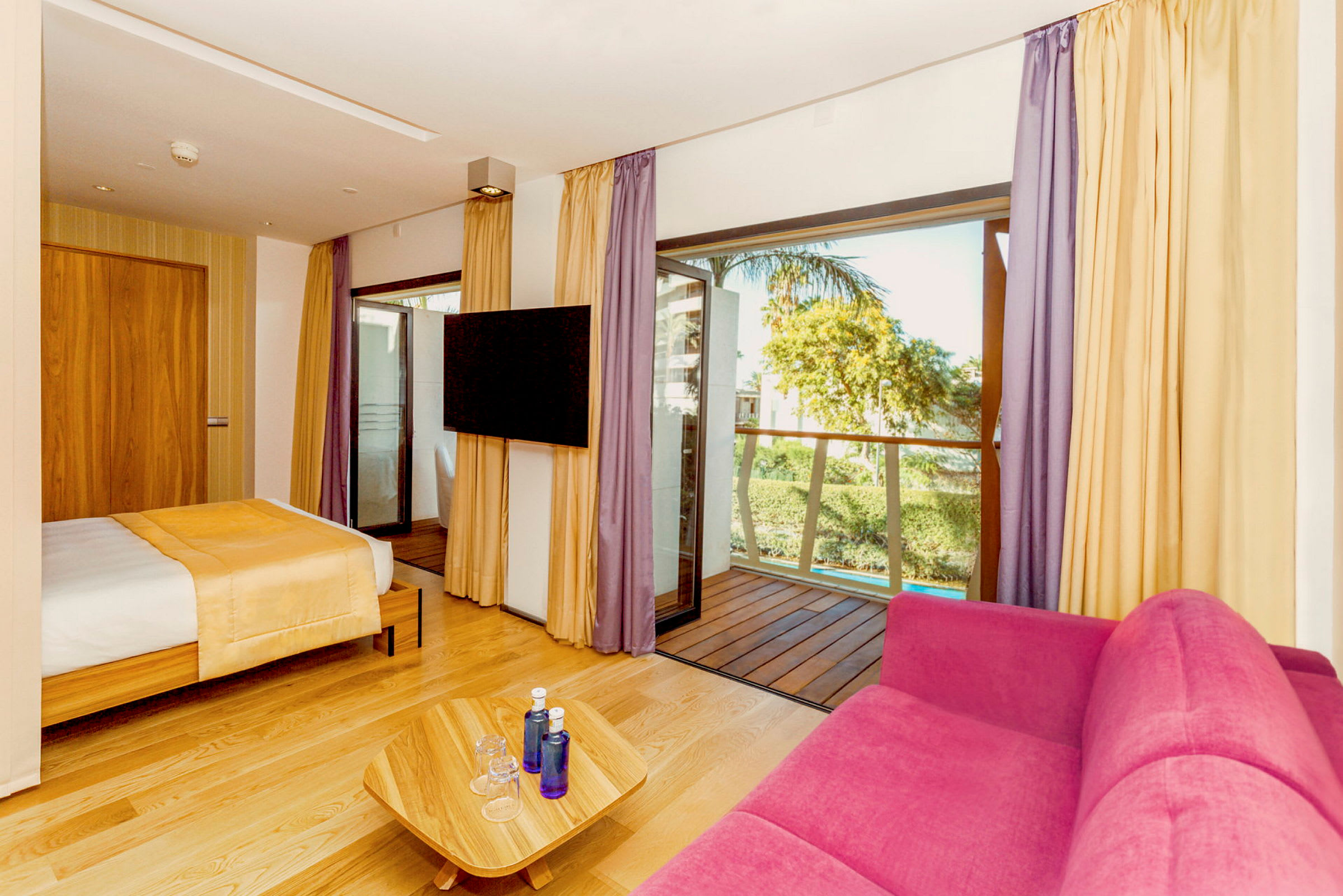 Hotelværelse med seng, pink sofa, TV og altan. (AI-genereret tekst)