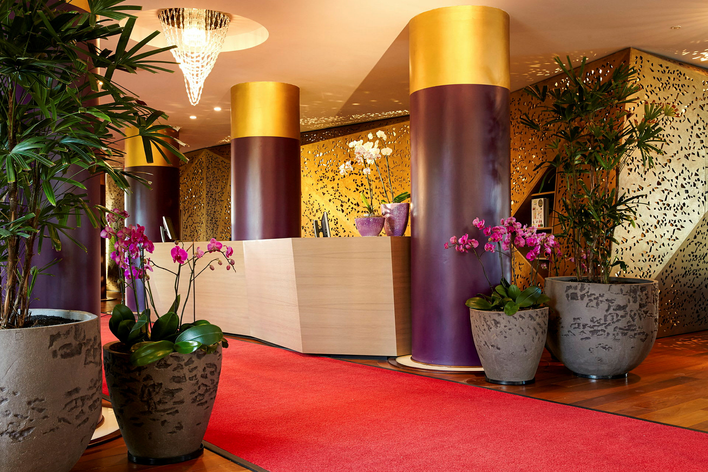 Receptionen i et elegant hotel med røde tæpper, store potteplanter og dekorative søjler. (AI-genereret tekst)