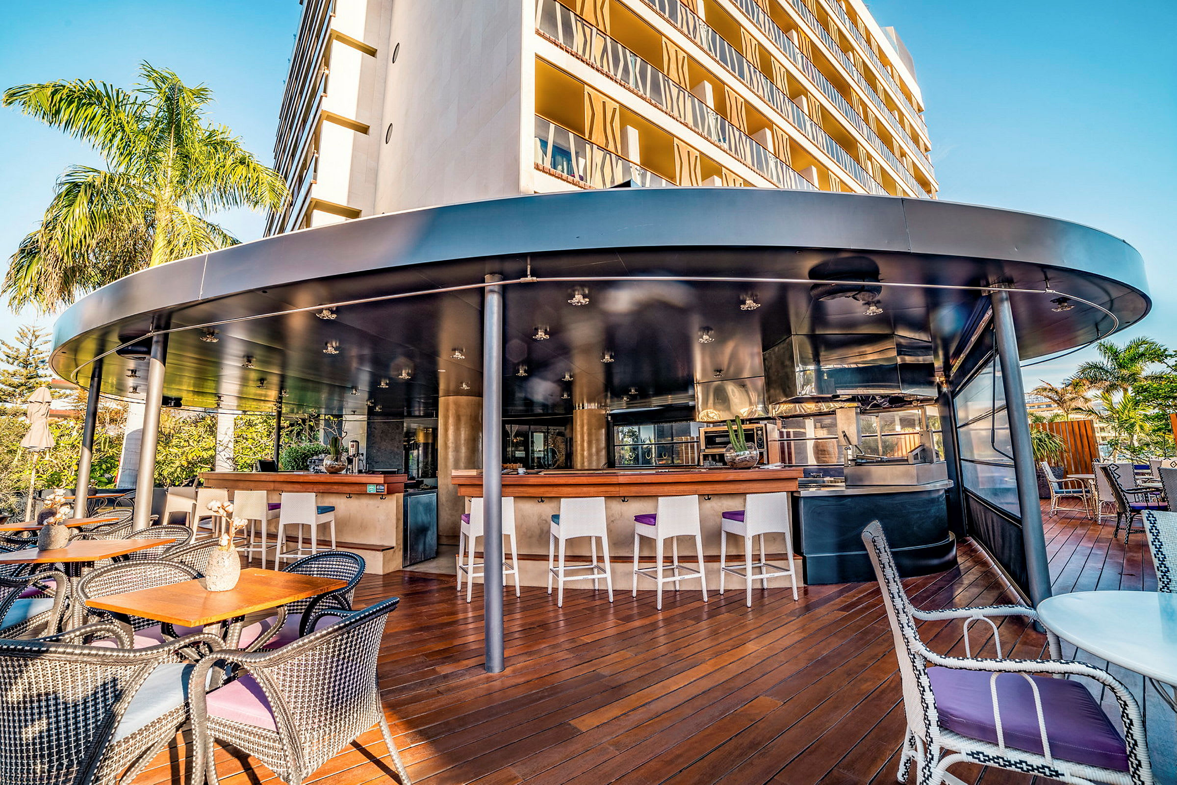 Et udendørs serveringsområde med borde, stole og en bar med høje stole under et rundt loft. En moderne bygning og palmer ses i baggrunden. (AI-genereret tekst)