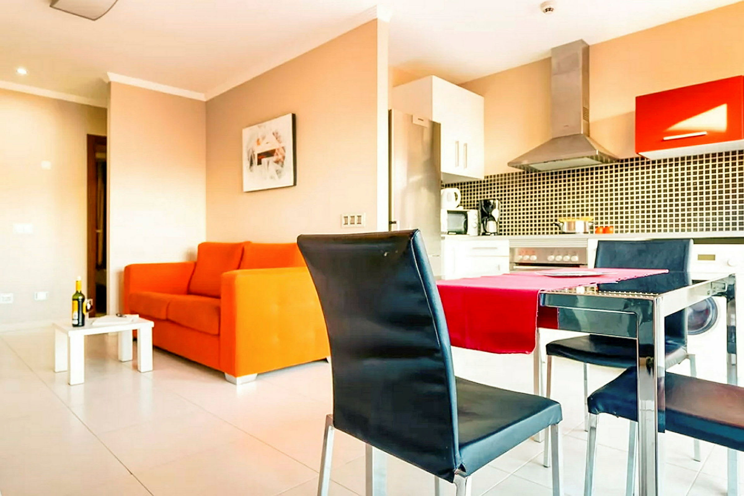 Stue med orange sofa, spisebord og stole, samt integreret køkken med komfur og køkkenudstyr. (AI-genereret tekst)