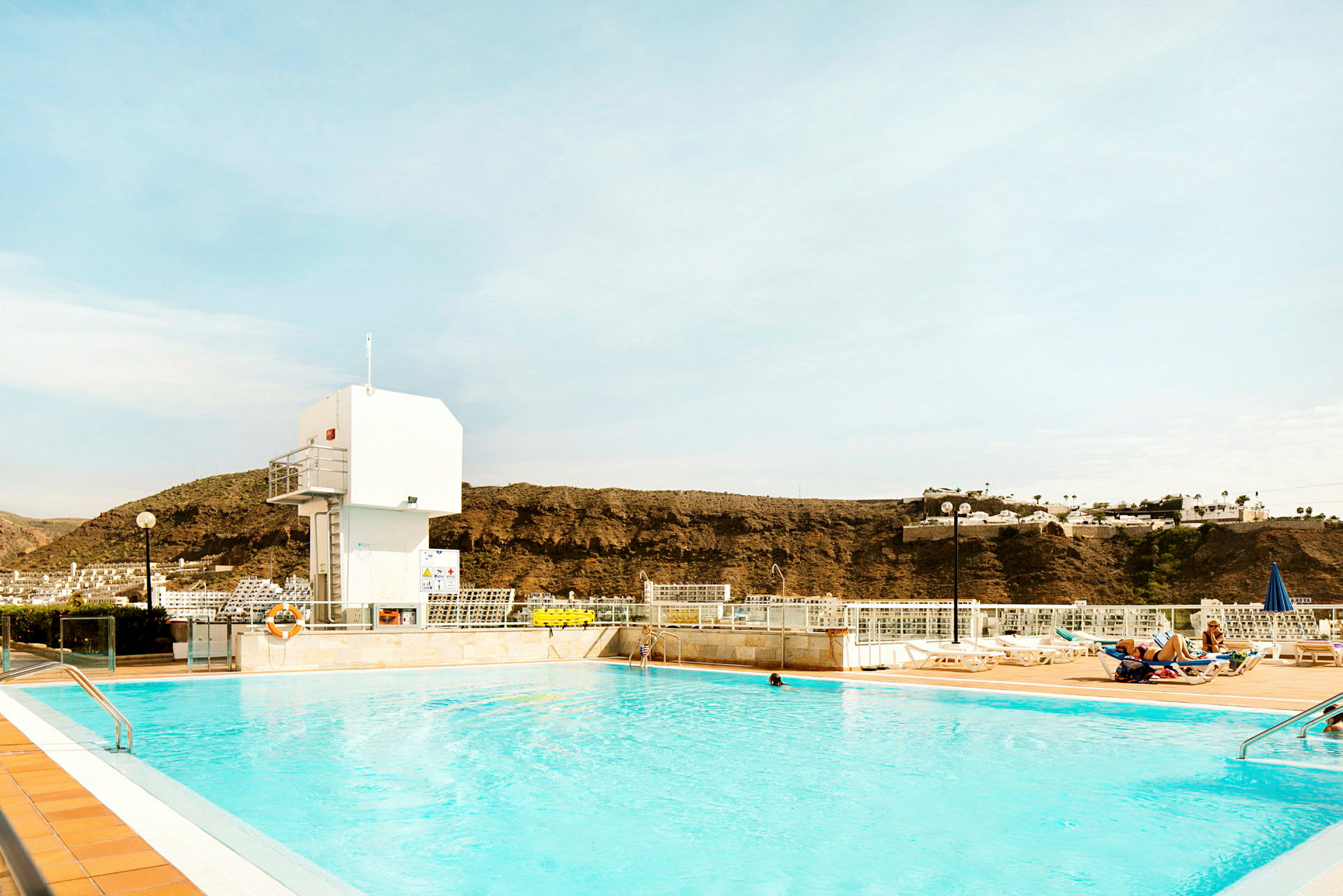 Udendørs pool med liggestole i baggrunden og bjerge i det fjerne. (AI-genereret tekst)