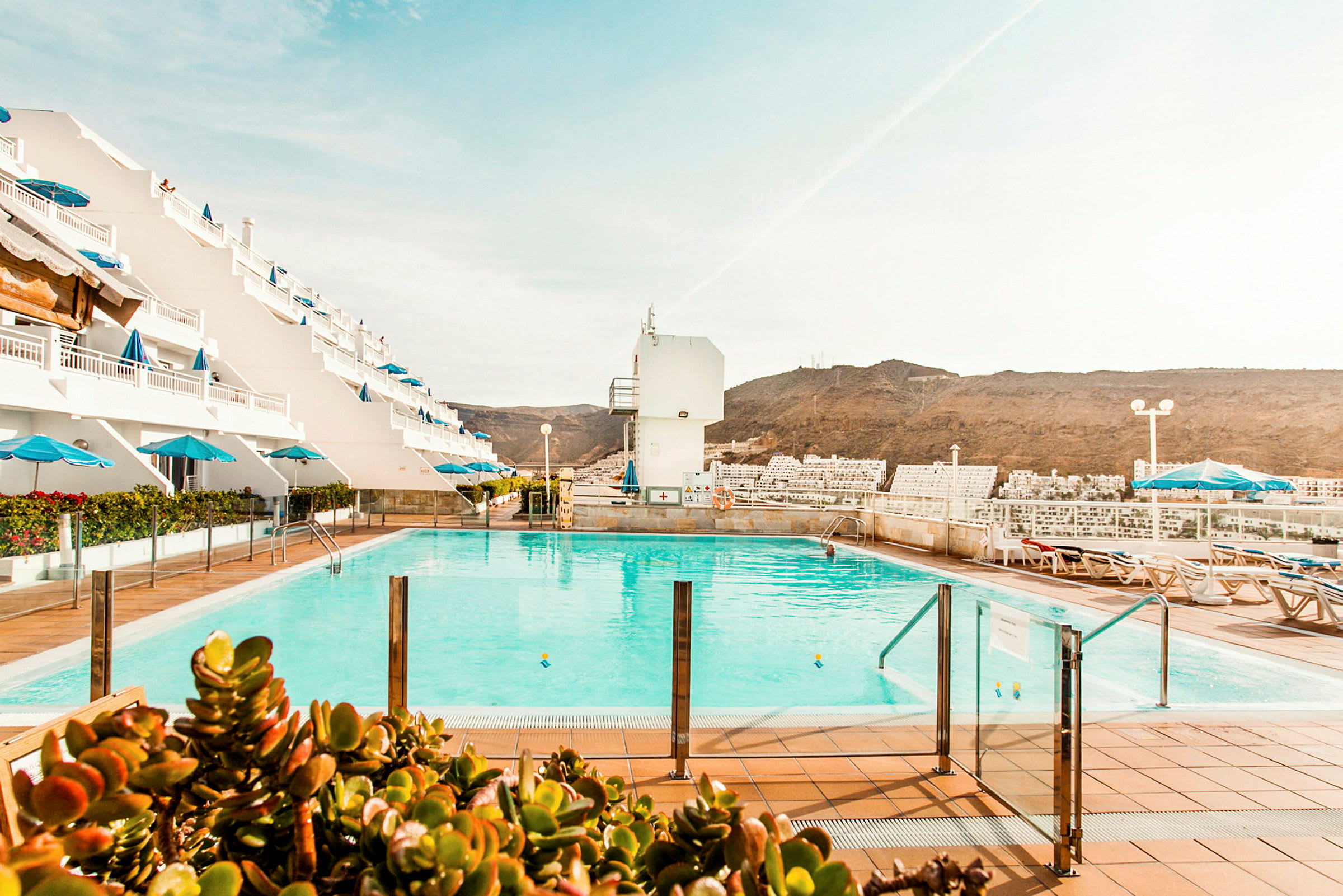 Poolområde med liggestole og parasoller, omgivet af et hotel med hvide facader og udsigt til bjergene. (AI-genereret tekst)