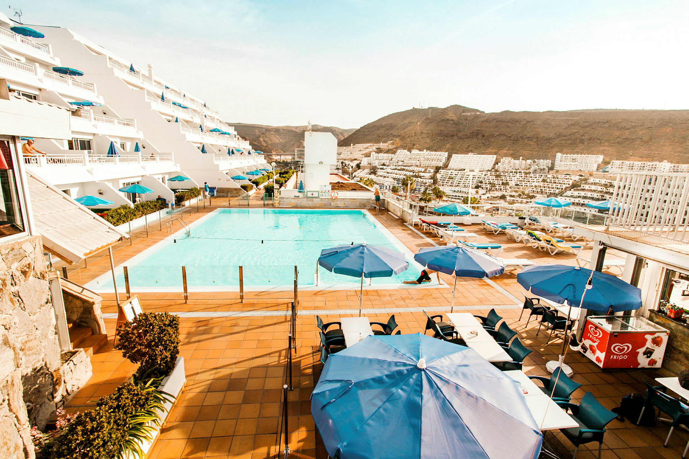 Billede af et anlæg med pool, liggestole og blå parasoller foran et hotel med terrasser. (AI-genereret tekst)