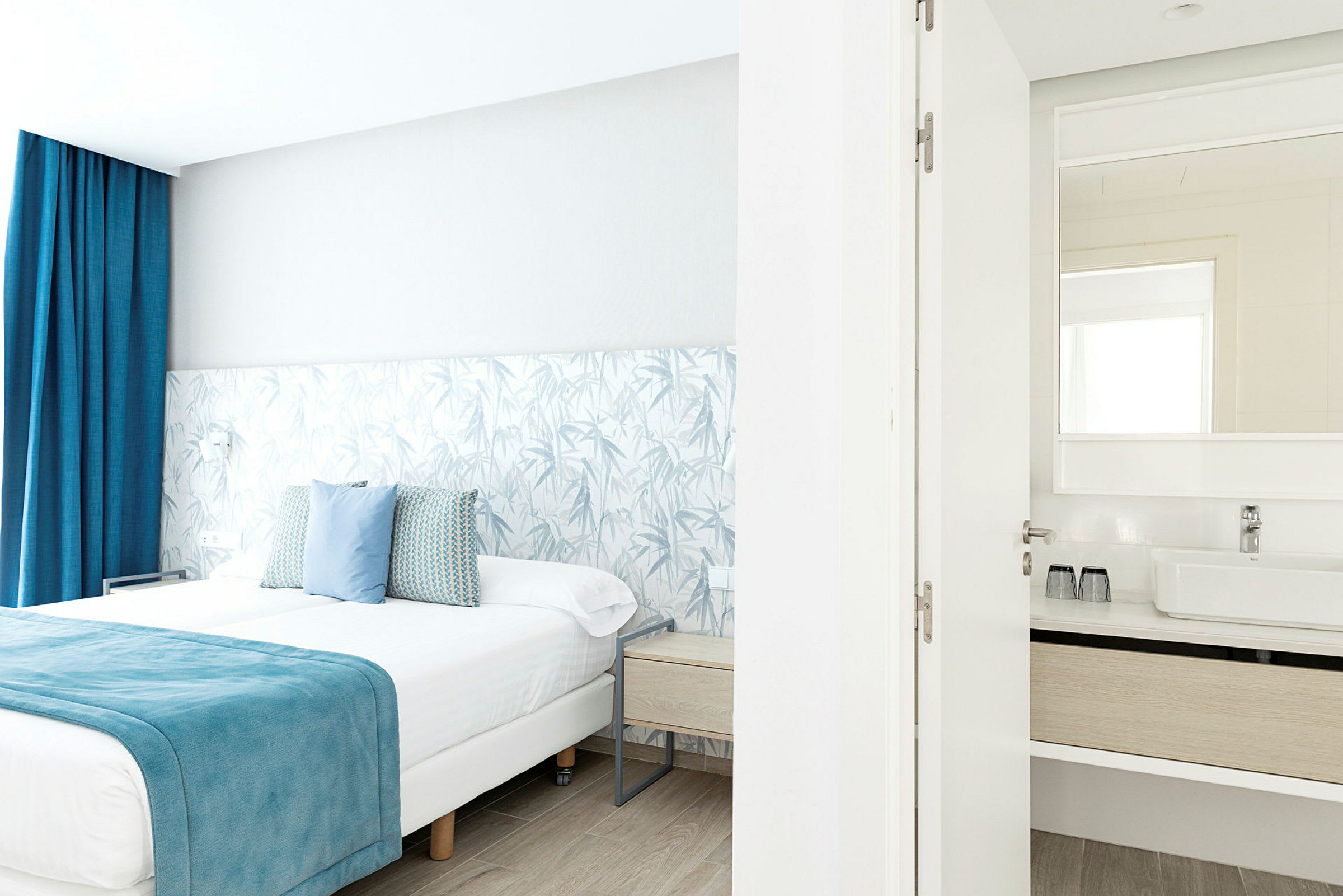 Et lyst hotelværelse med en dobbeltseng med blå detaljer og et tilknyttet badeværelse med synligt spejl. (AI-genereret tekst)