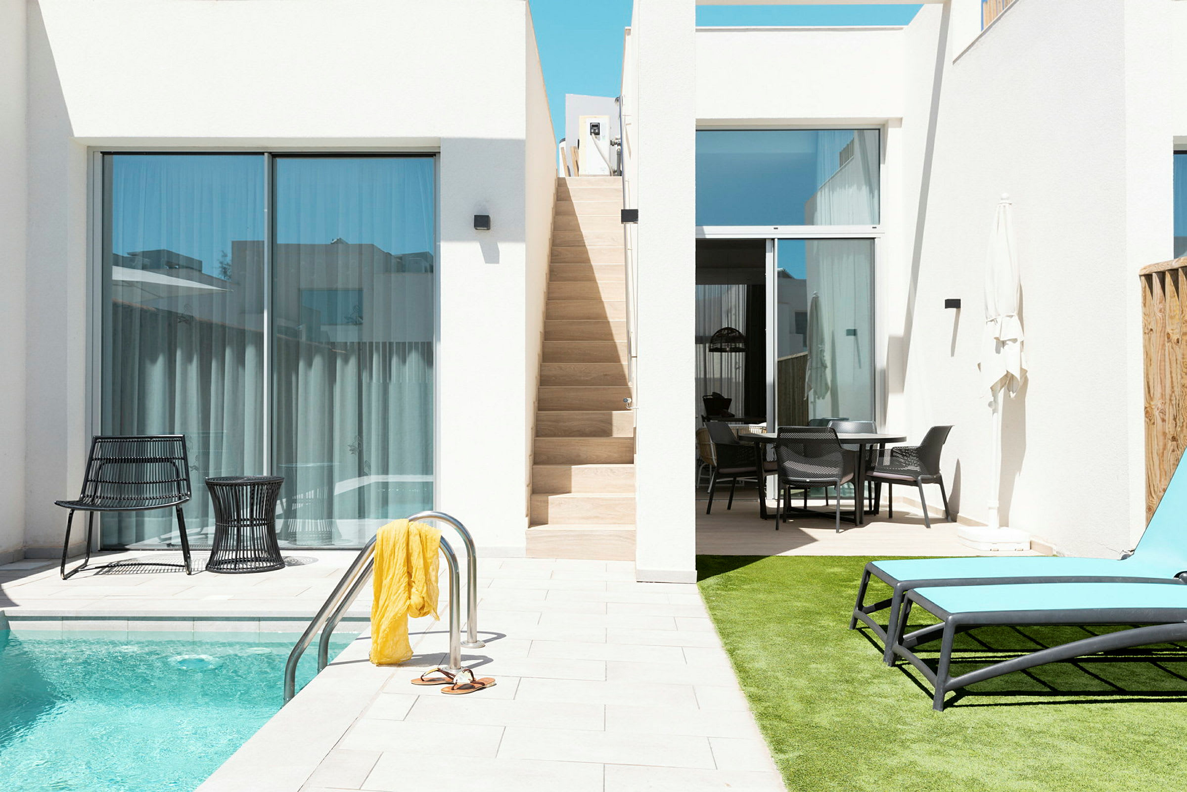 En pool med liggestole og et bord med stole ved siden af, samt en trappe der fører op til en balkon. (AI-genereret tekst)
